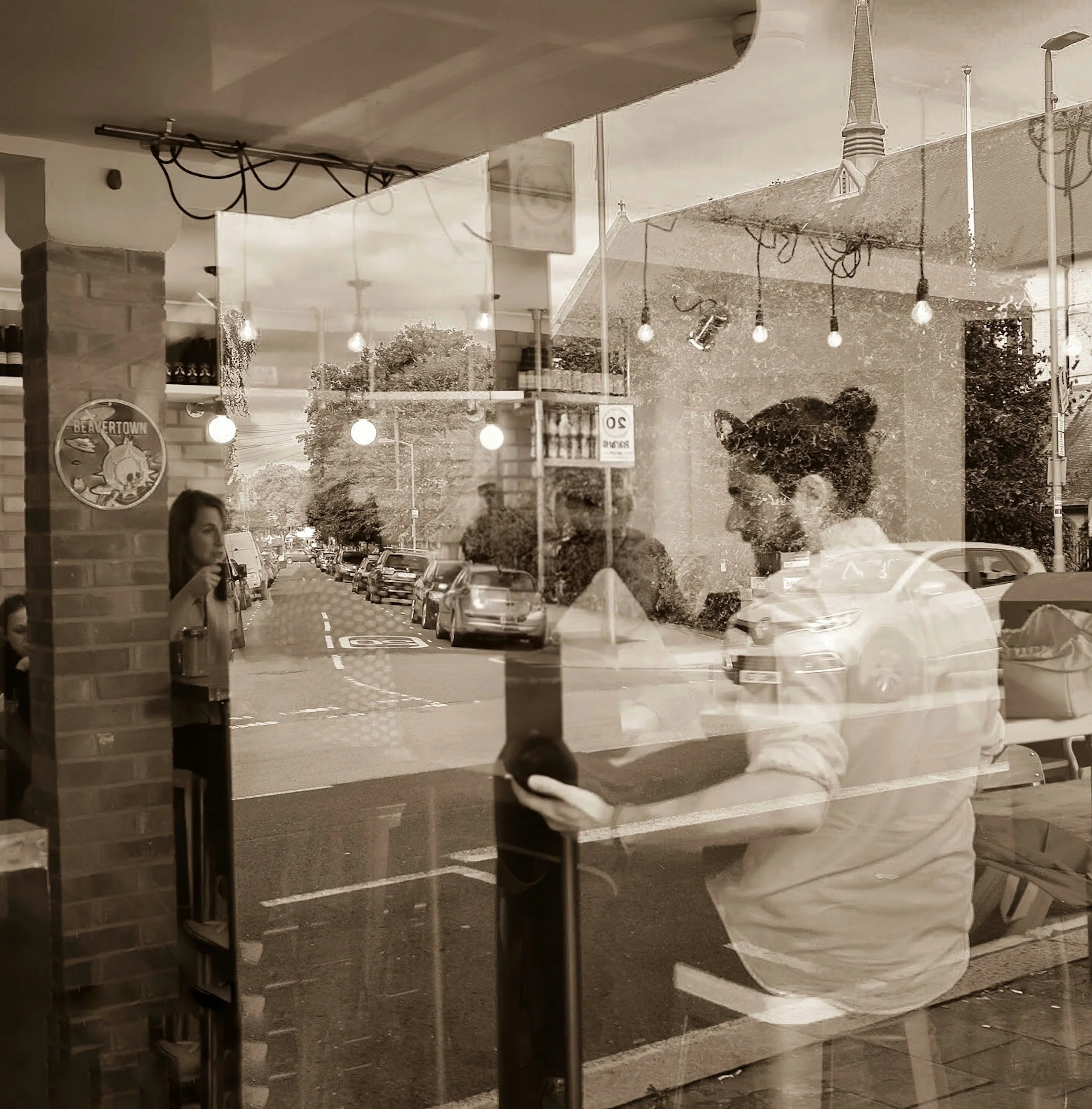 Zaid, one of the founders, inside an artisan coffee café with a coffee cup, looking through the large glass window at the street outside where parked cars and some trees can be seen.