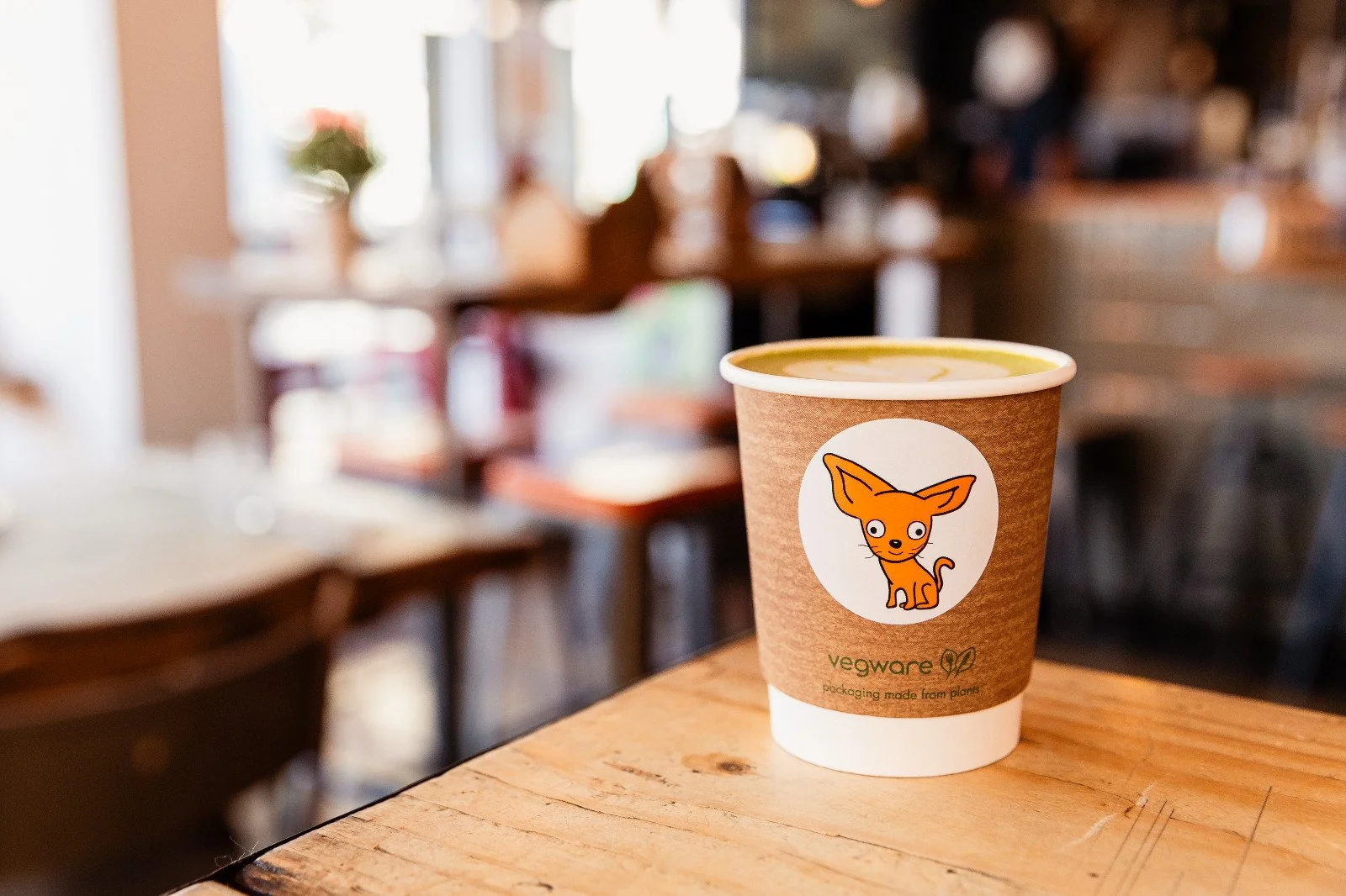 A brown paper cup with a cartoon fox logo and the text 'vegware packaging made from plants' on a wooden table in a cozy cafe with blurred background. Specialty coffee and vibrant brunch dishes.