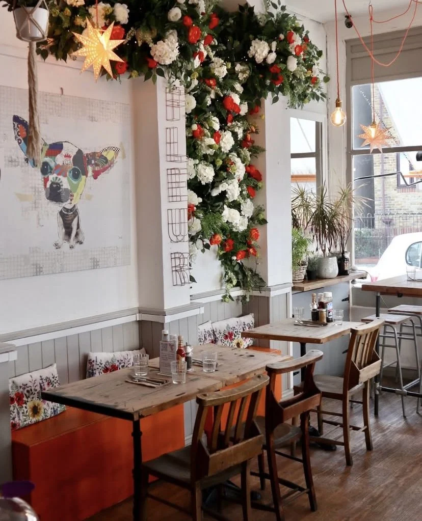 A neighbourhood café serving vibrant, healthy brunch. An interior with wooden tables and chairs, decorated with floral cushions. A large floral arrangement with white and red flowers decorates a wall corner.