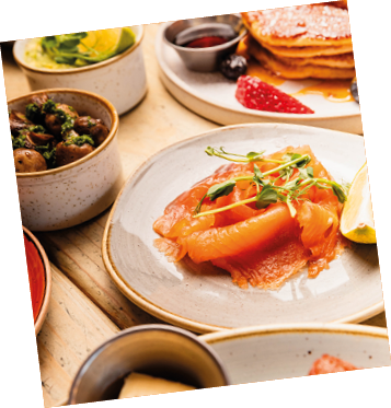 Salmon with microgreens and lemon on a white plate, with pancakes, berries, and other breakfast items in the background. Middle Eastern and Mediterranean inspired breakfast and brunch