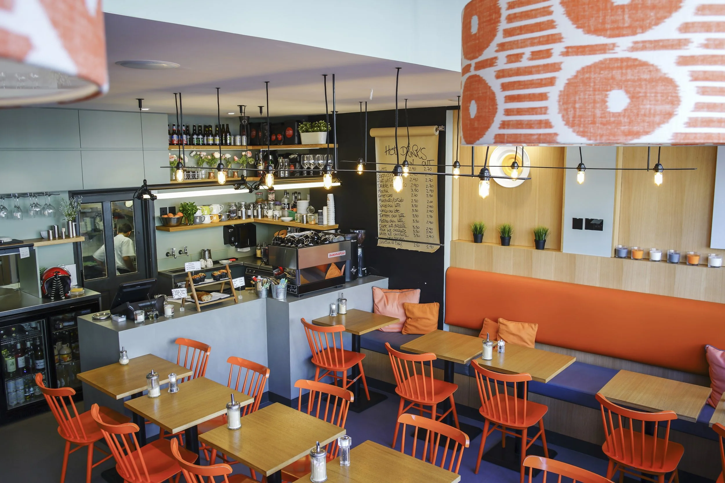 A local favourite for brunch in Wandsworth. Interior of Cafe Tamra, an independent brunch café with wooden tables and orange chairs, a barista station with coffee equipment, shelves with bottles and glasses, and a menu board.