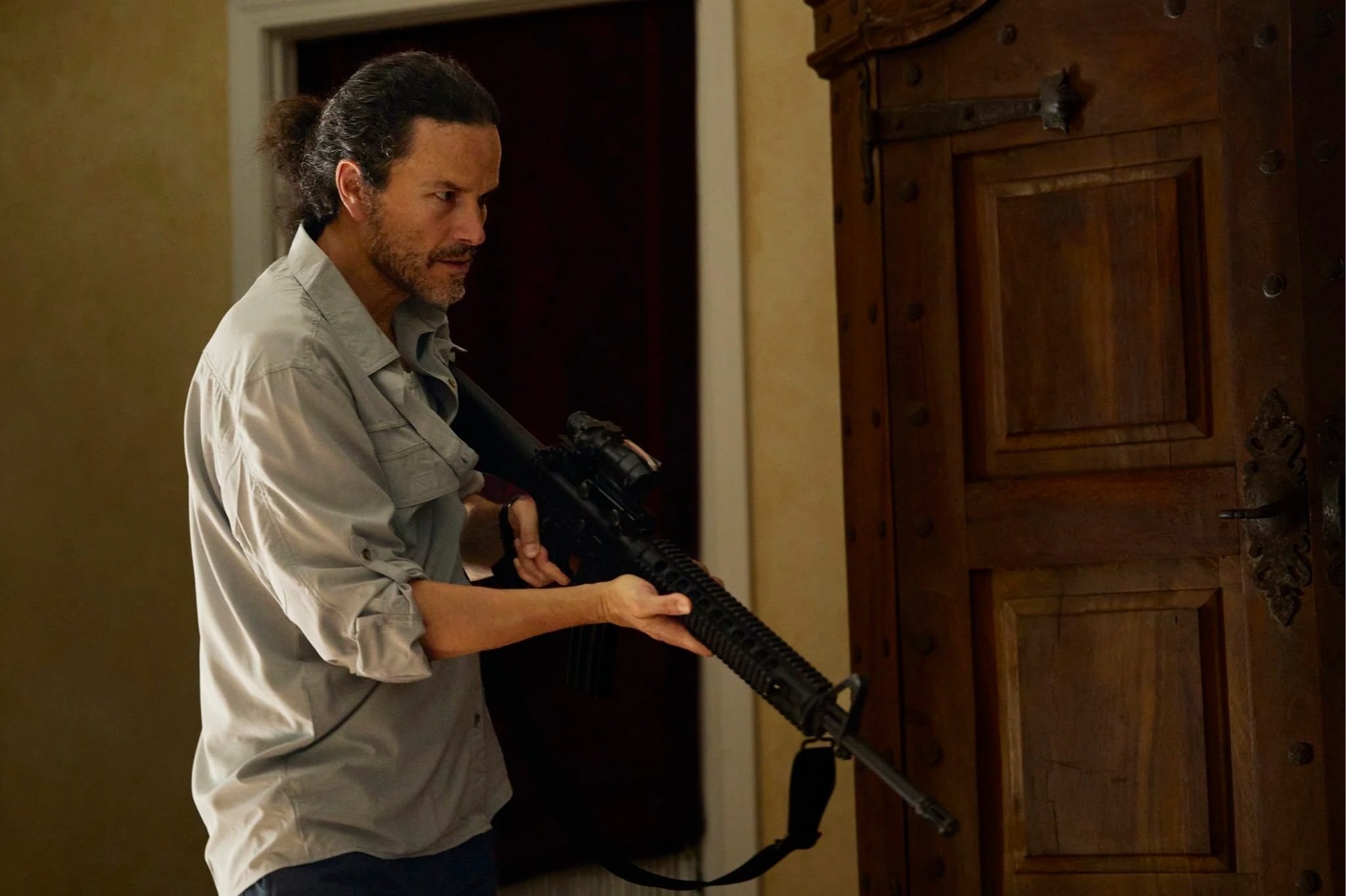 Professional unit still of a man with long, dark hair tied back, holding a rifle, standing in a room next to a wooden cabinet, for the short film "The Harvest."