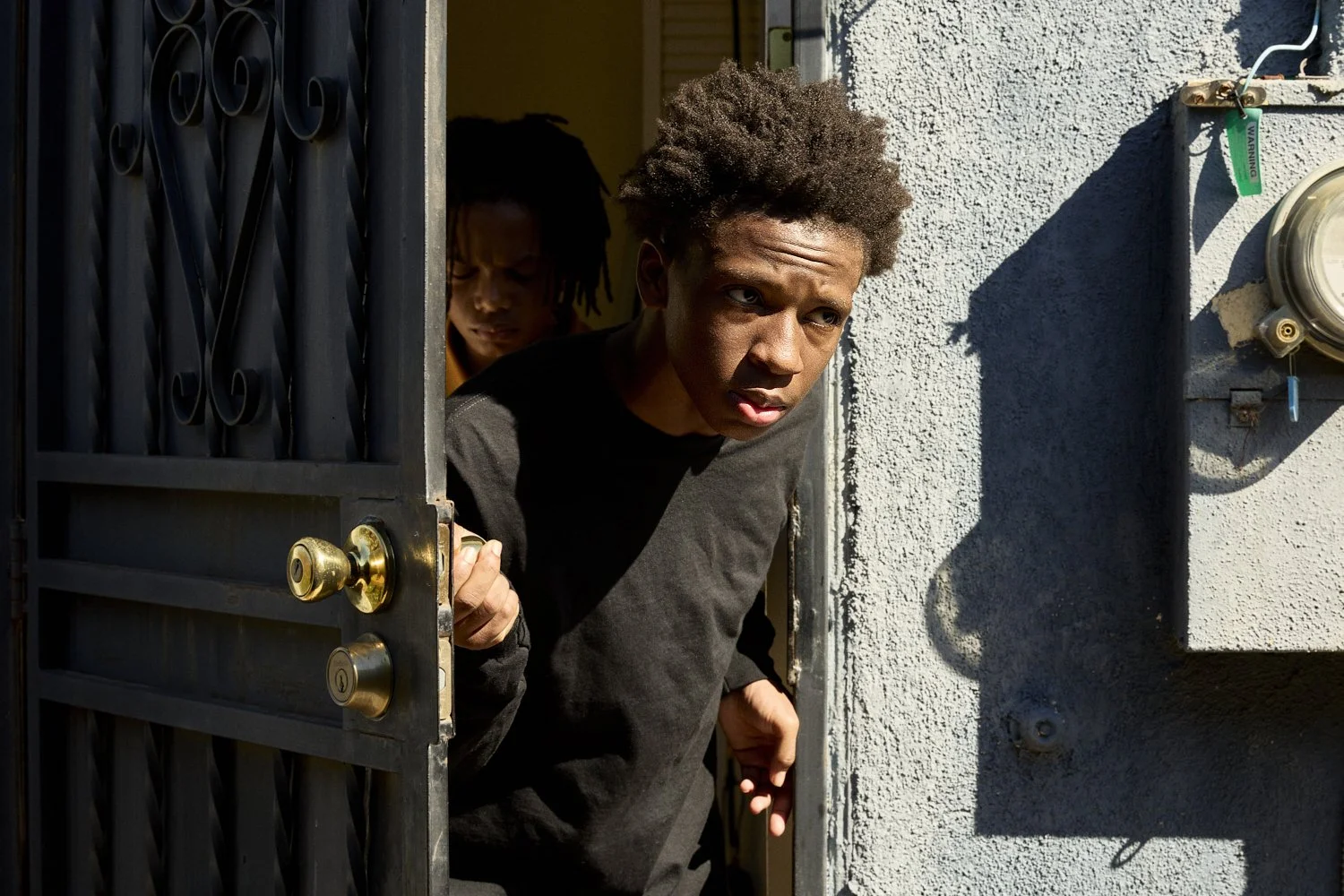 Professional film still of two young black men peeking out from behind a partially open gate outside a building on a sunny day for the short film "Now I Lay Me."
