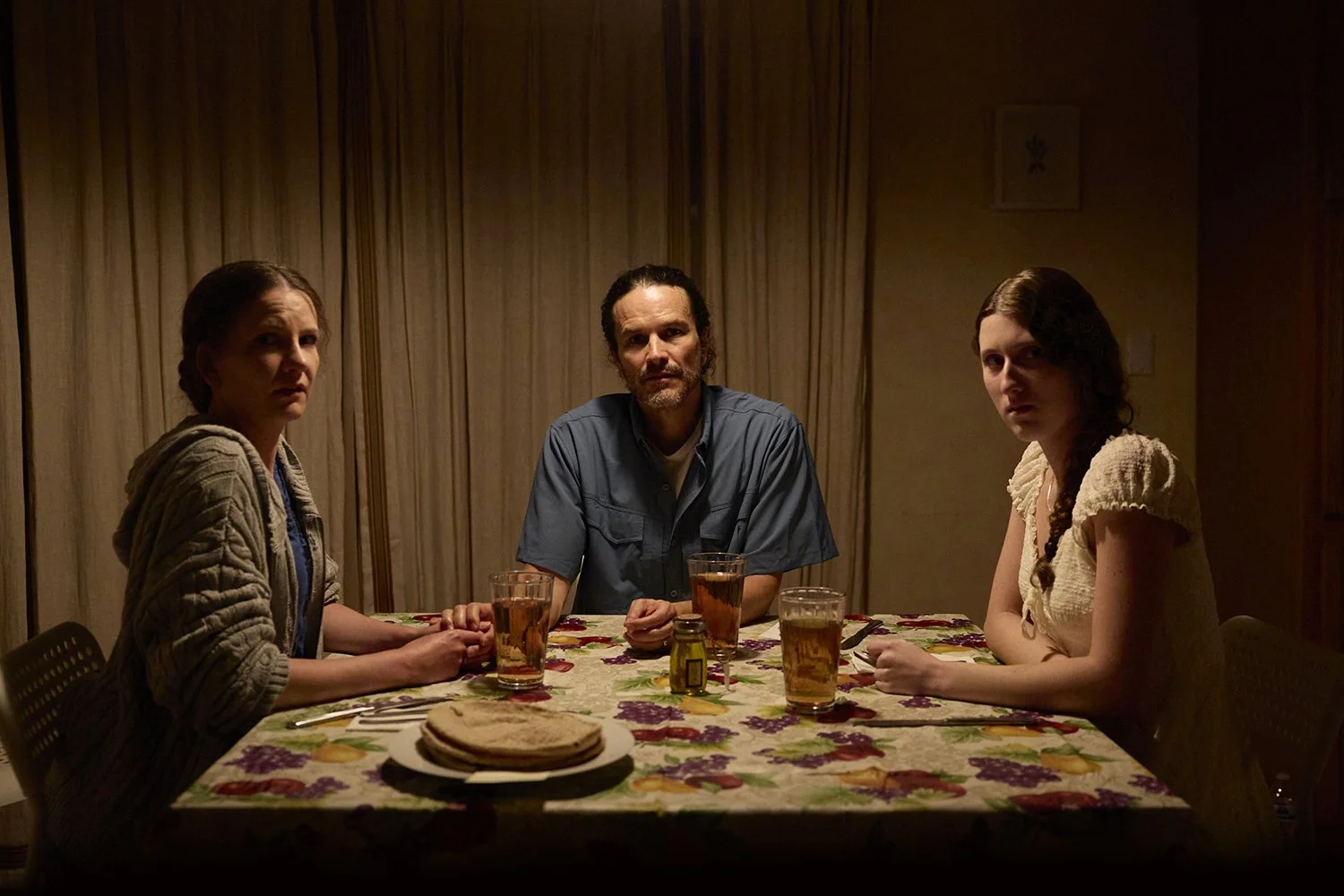 Professional film still of three people sitting at a dimly-lit dining table, looking at the camera, for the short film "The Harvest."