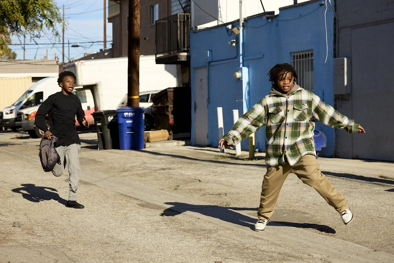 Professional unit stills of two teenage black boys are in an urban alleyway, one running with a backpack and the other walking with his arms outstretched, surrounded by parked cars and blue walls for the short film "Now You Lay Me."