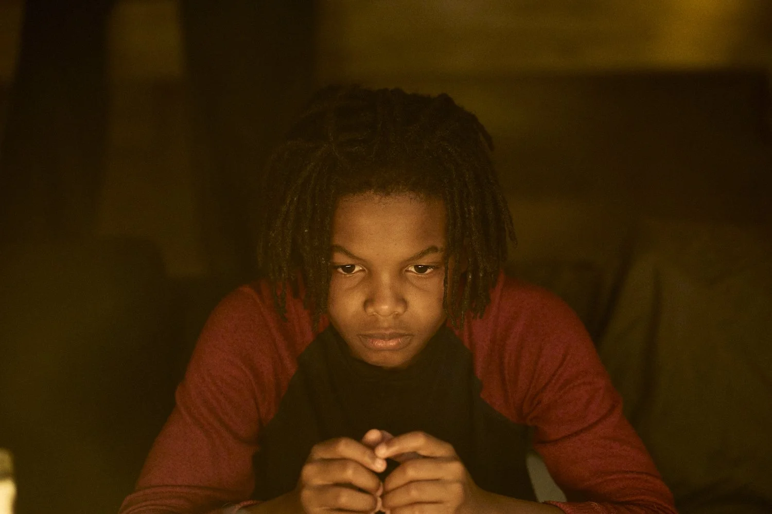 Professional unit still of a young black man with dreadlocks and a red and black shirt, sitting at a table in a dimly lit room, looking down with hands clasped for the short film "Now You Lay me."