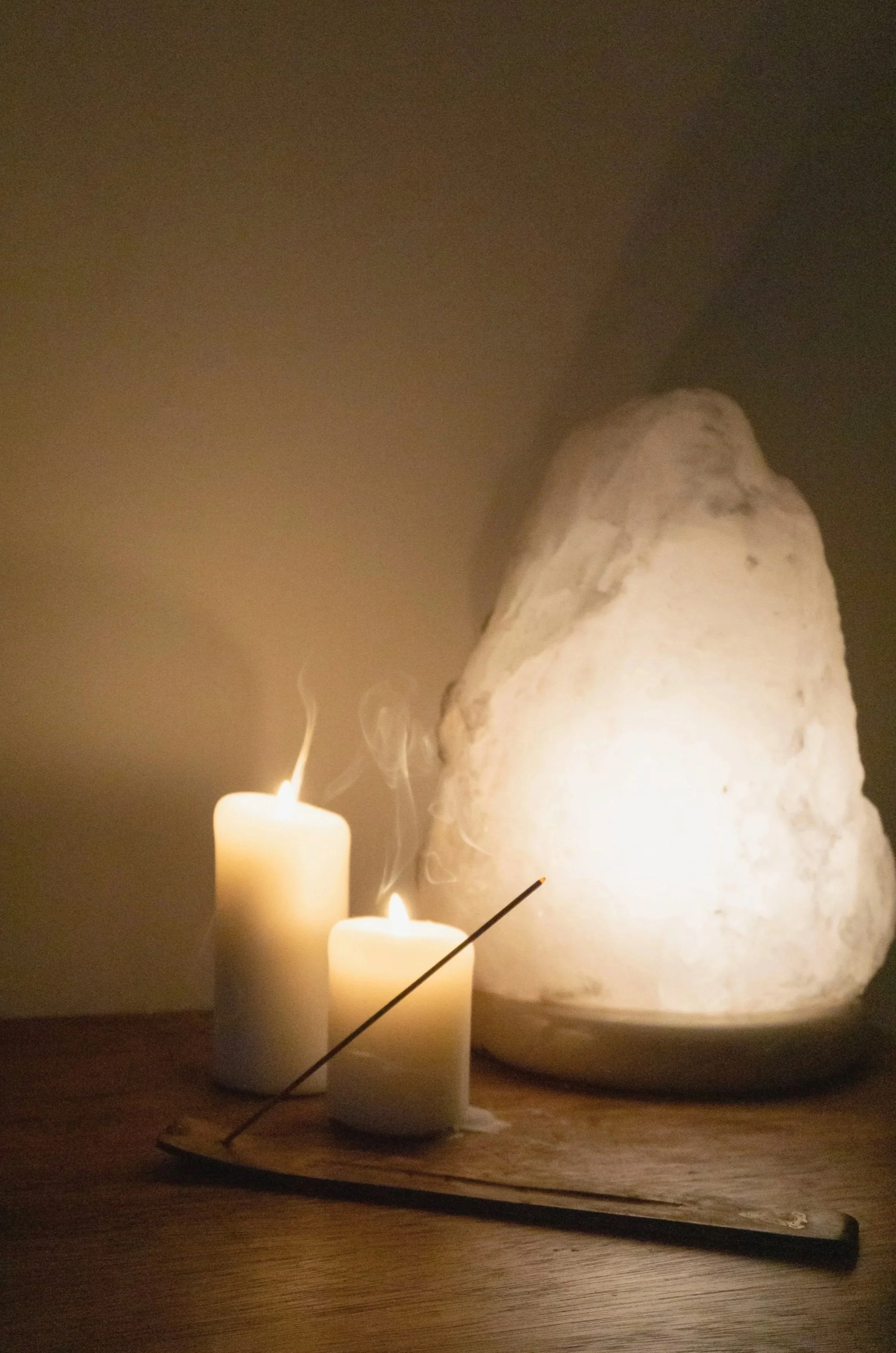 A set of three white candles of varying heights on a wooden surface, with a lit matchstick near the candles. In the background, there's a large, irregularly shaped white rock with a rough surface, illuminated softly, creating a warm, cozy atmosphere.