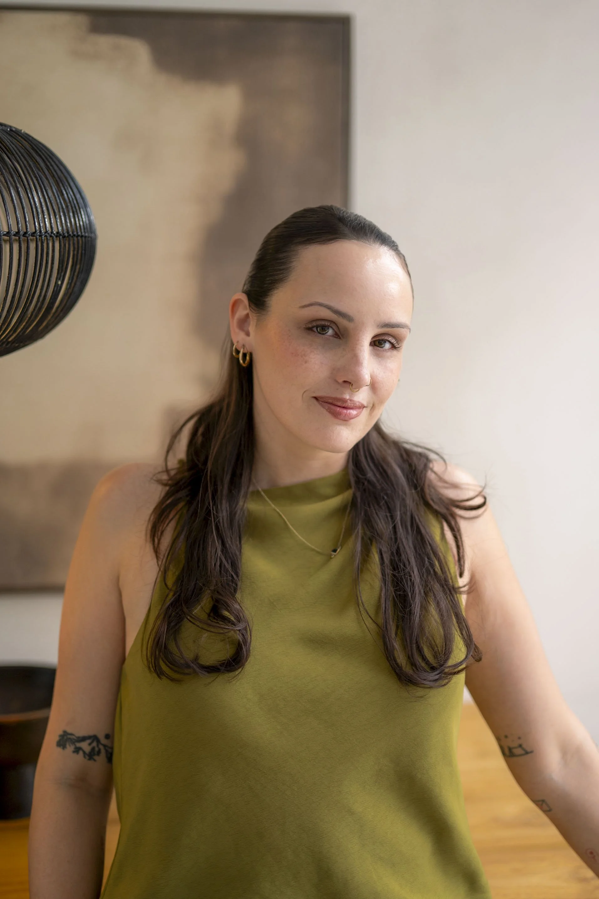 A woman with long, wavy brown hair wearing a sleeveless, olive green top, gold hoop earrings, and small tattoos on her arms, smiling in a room with neutral-colored walls and a black fan.