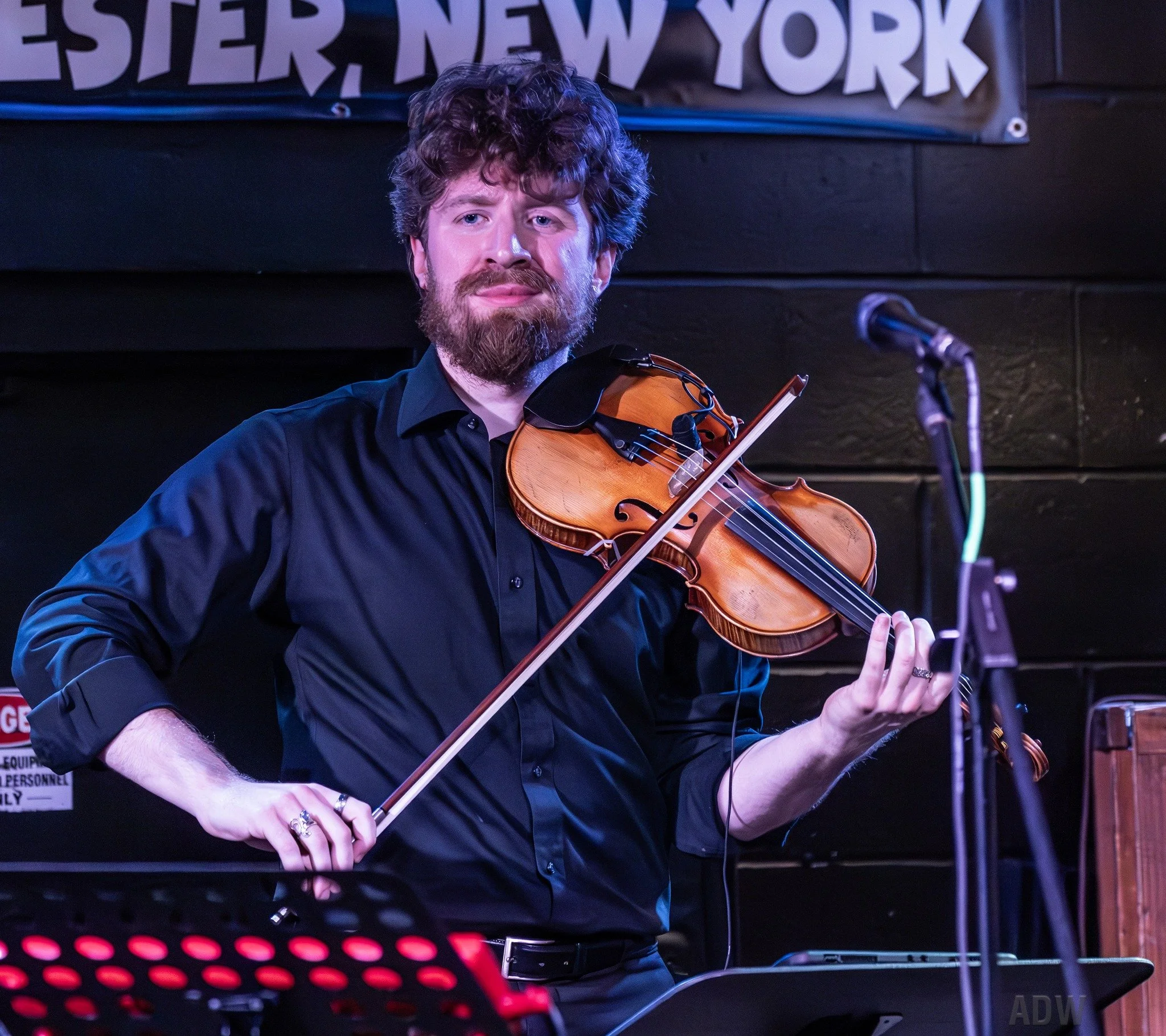 A man with curly hair and a beard playing a violin onstage against a black background.