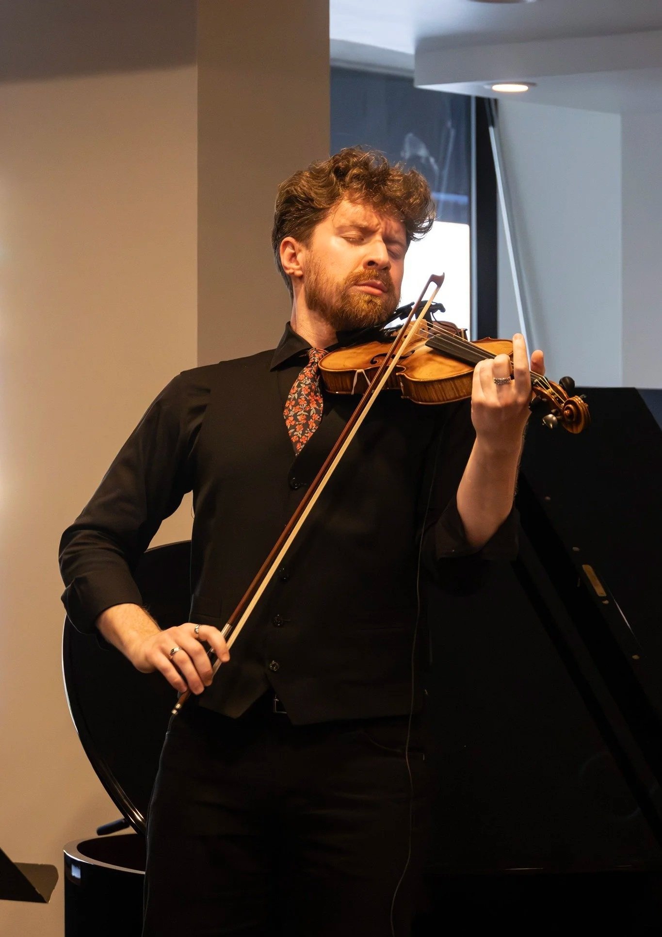 A man in black clothing playing a violin in a room with a black grand piano in the background.