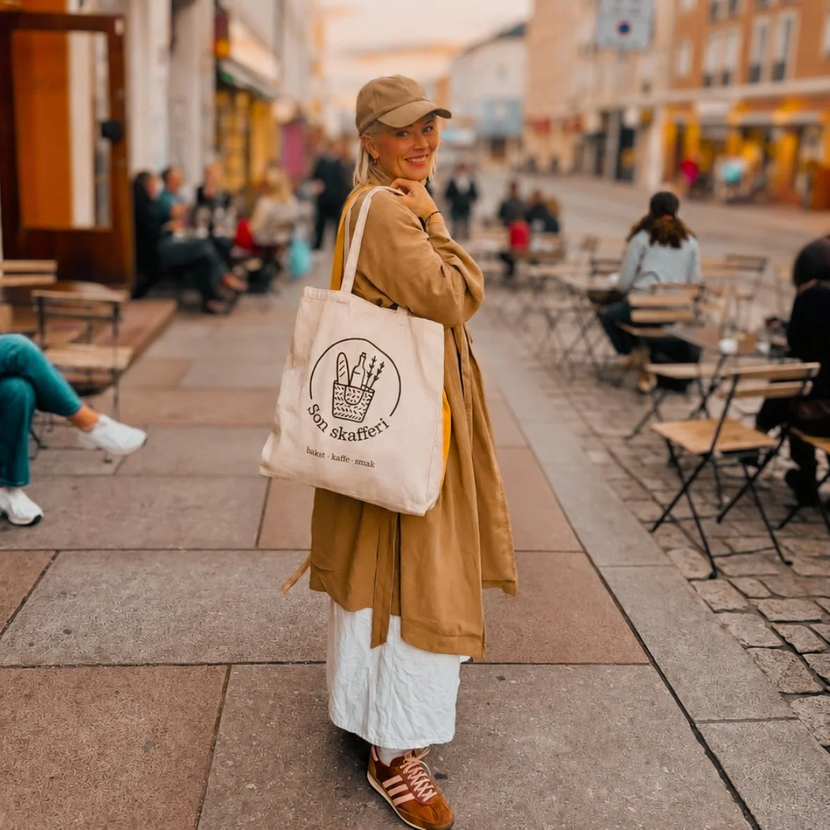 Smilende kvinne i beige frakk og caps med handlenett fra «Son Skaffer» på en travel kafégate.