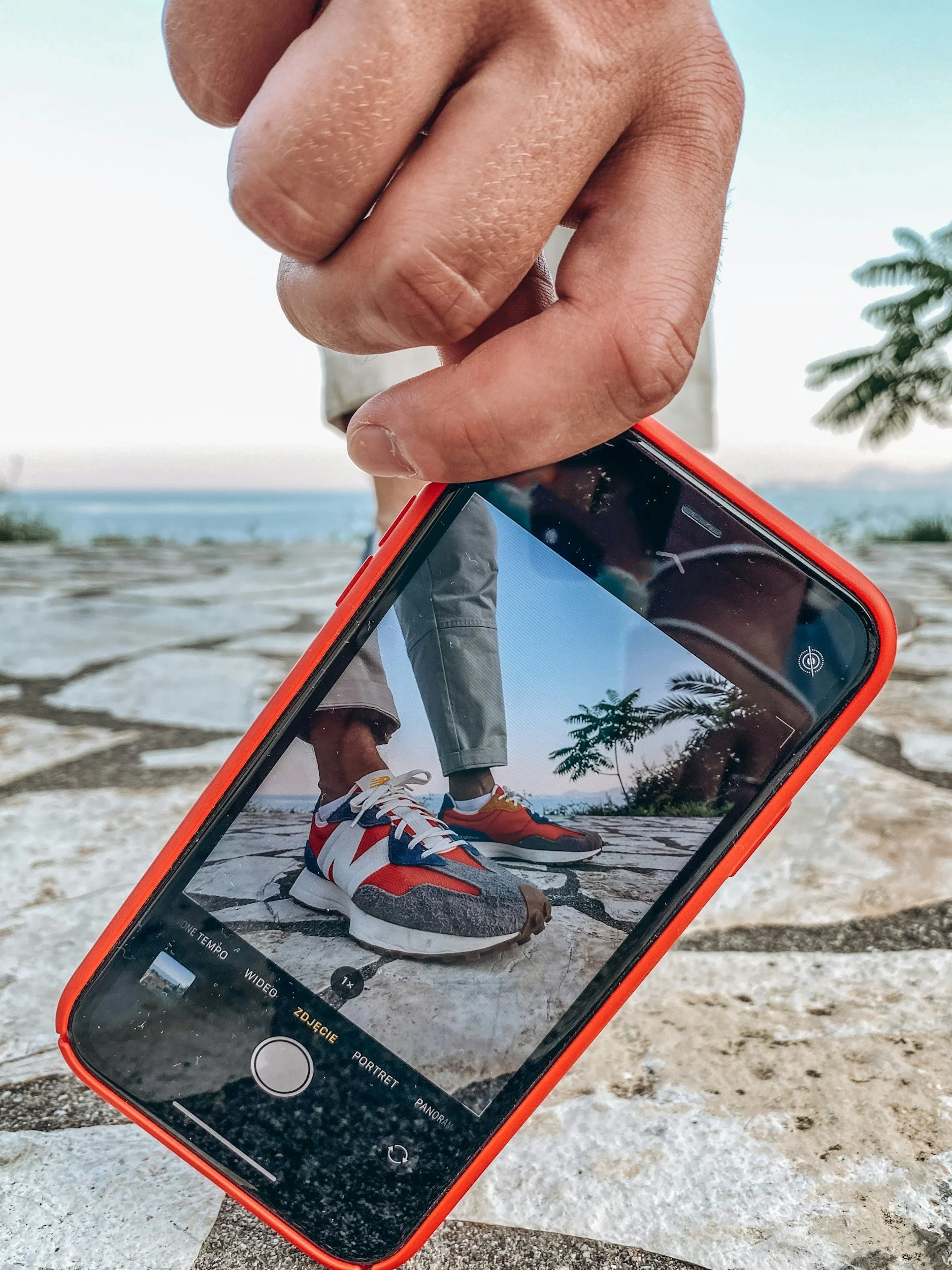 Een hand houdt een smartphone vast die een foto van een persoon in sneakers en sportkleding weergeeft, terwijl die persoon op een stenen vloer staat met een strand en zee op de achtergrond.