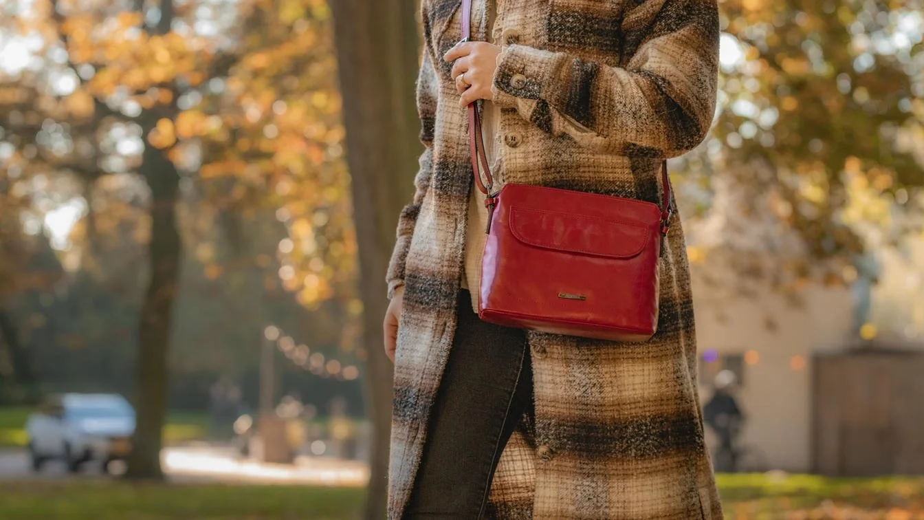 Een persoon draagt een rood schoudertasje en een wollen jas met een bruine ruitpatroon, in een herfstpark met gekleurd blad.