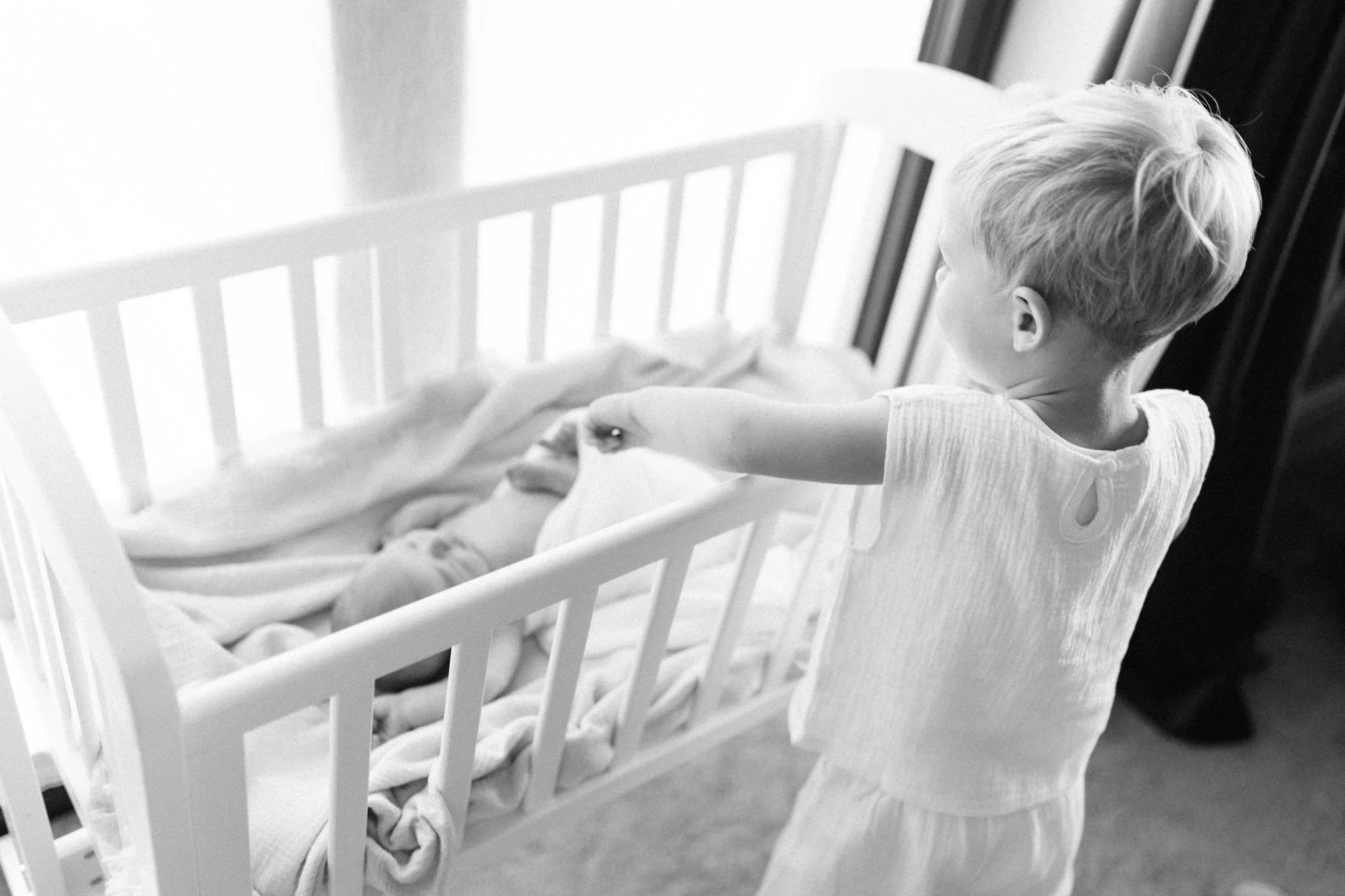 Toddler inspects newborn baby sister in her crib during a natural at-home newborn session in Cornwall