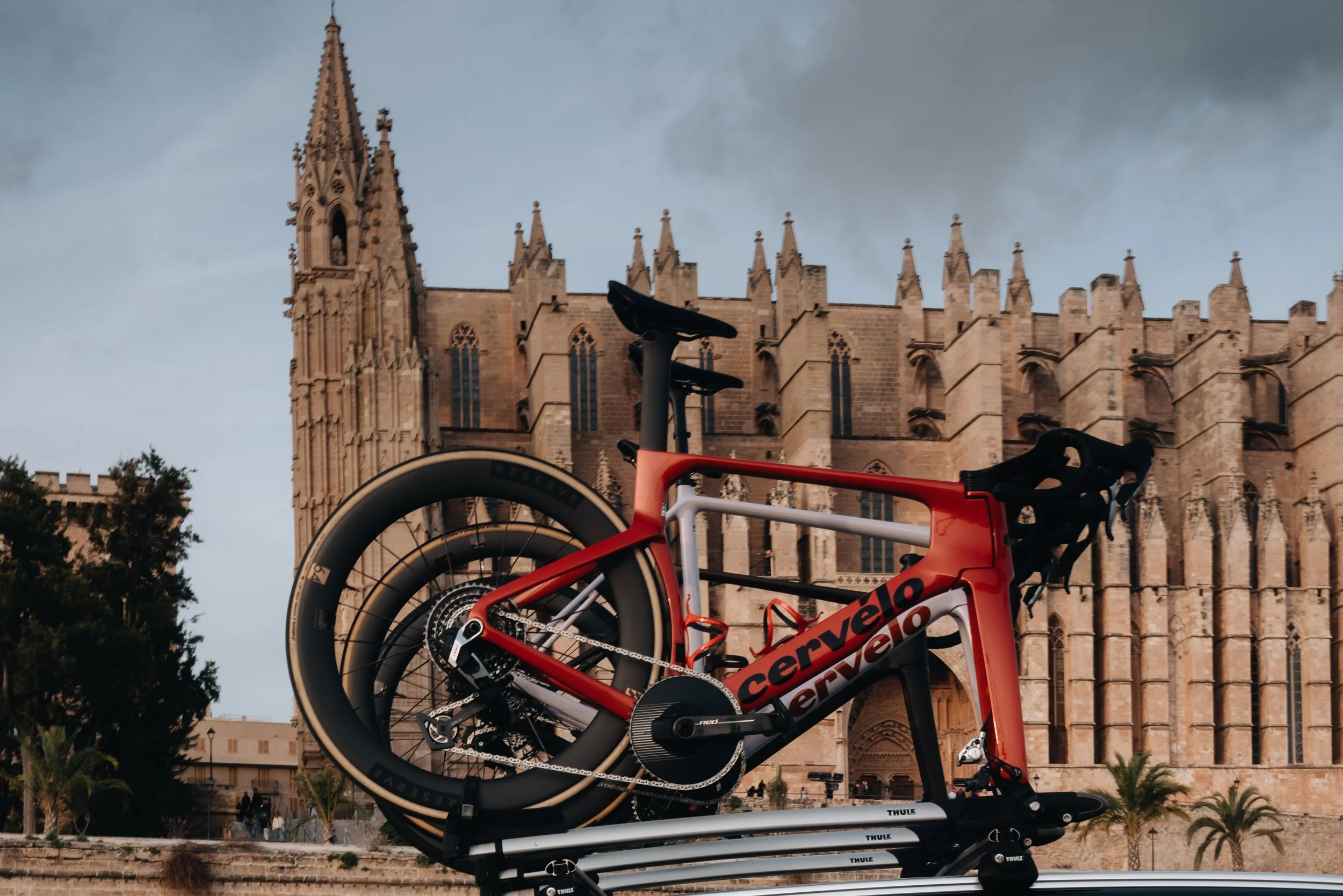 A red and black Cervelo road bicycle mounted on a roof rack in front of a Gothic cathedral with tall spires and intricate stonework.