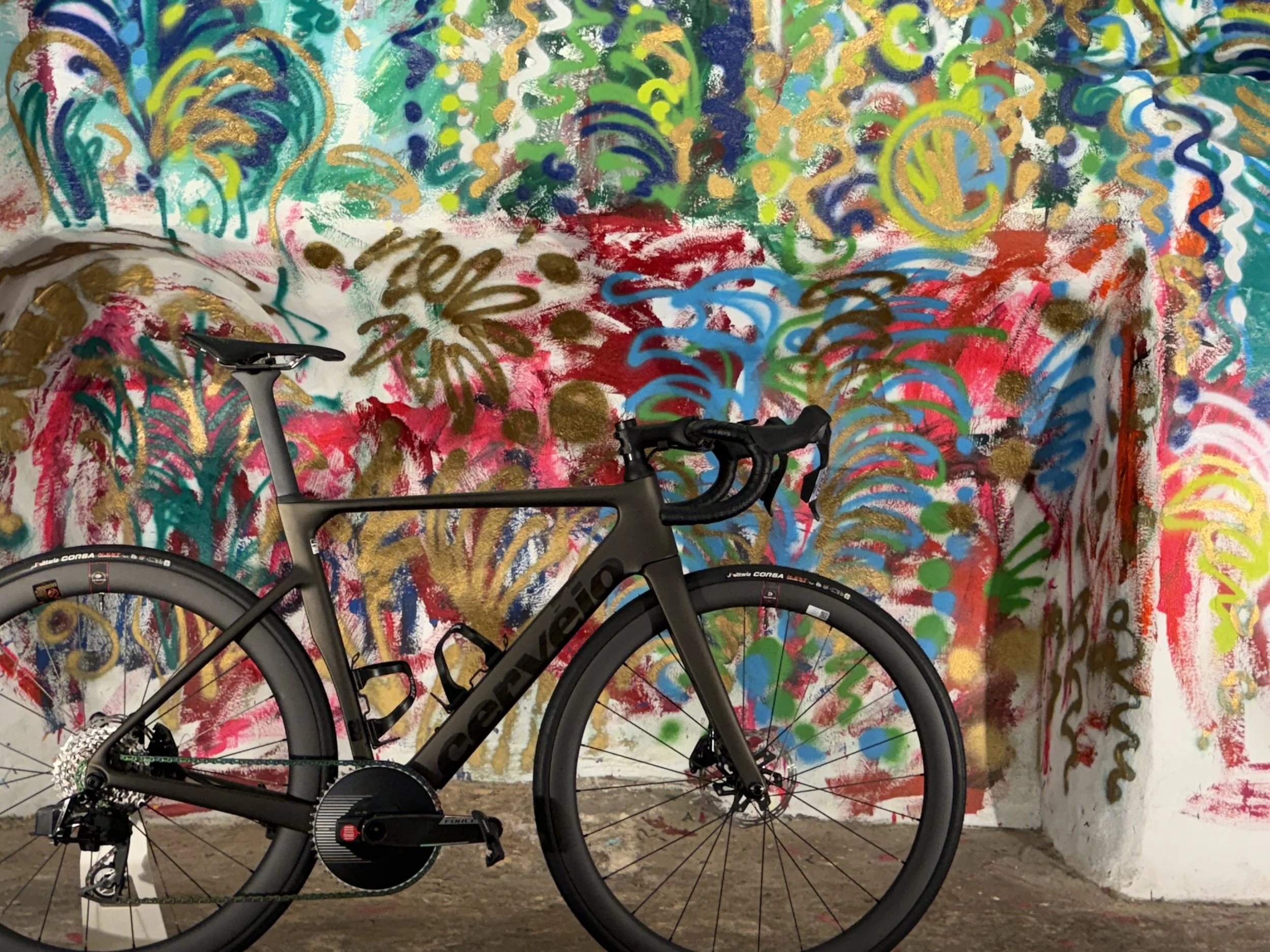 A black road bicycle with a matte finish frame is parked in front of a colorful, abstract graffiti wall with swirls, dots, and strokes in various bright colors.
