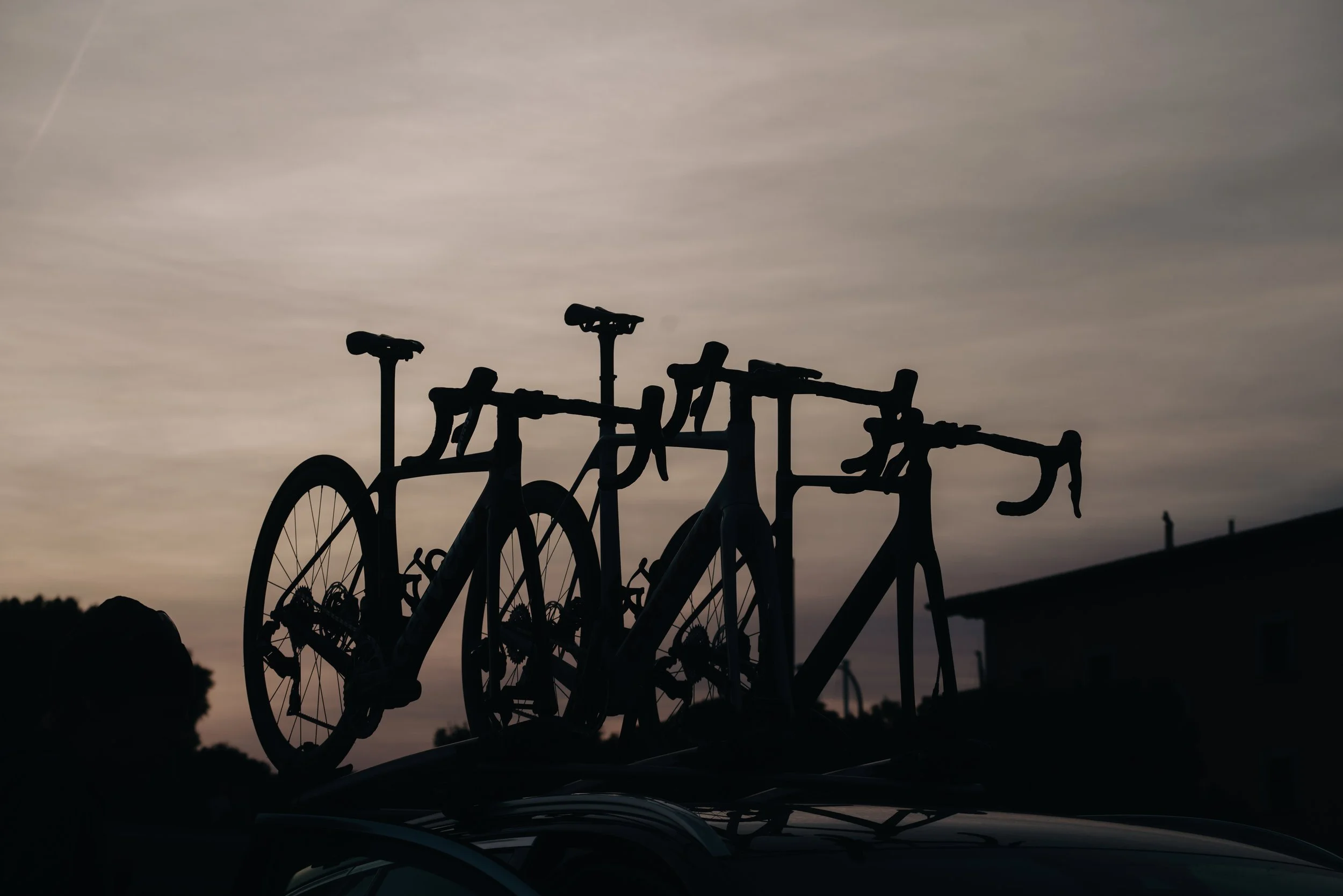 Silhouette of three bicycles mounted on top of a car against a cloudy sky at dusk.