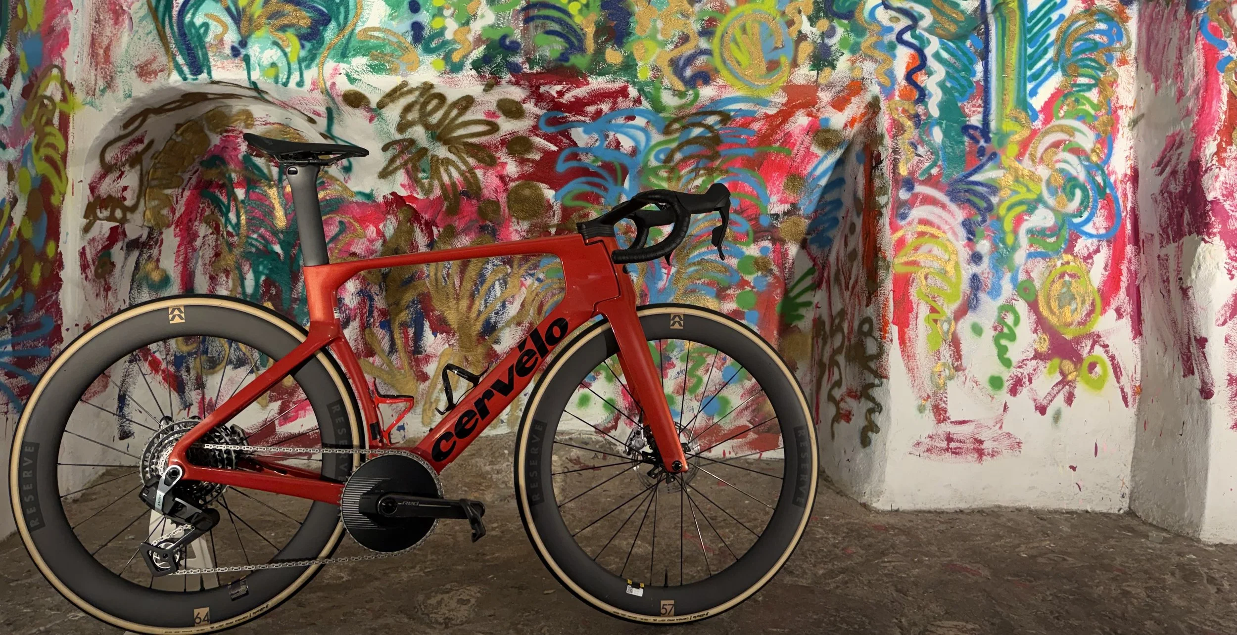 Red Cervelo road bike with black and tan tires in front of a colorful, abstract graffiti wall