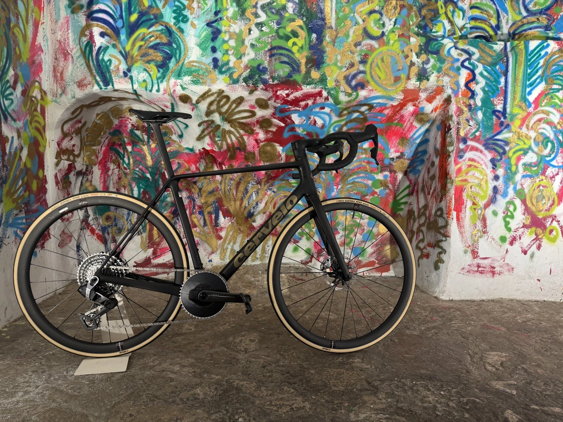 A black Cervélo road bicycle with beige tires is positioned in front of a colorful, abstract graffiti mural on a concrete wall.
