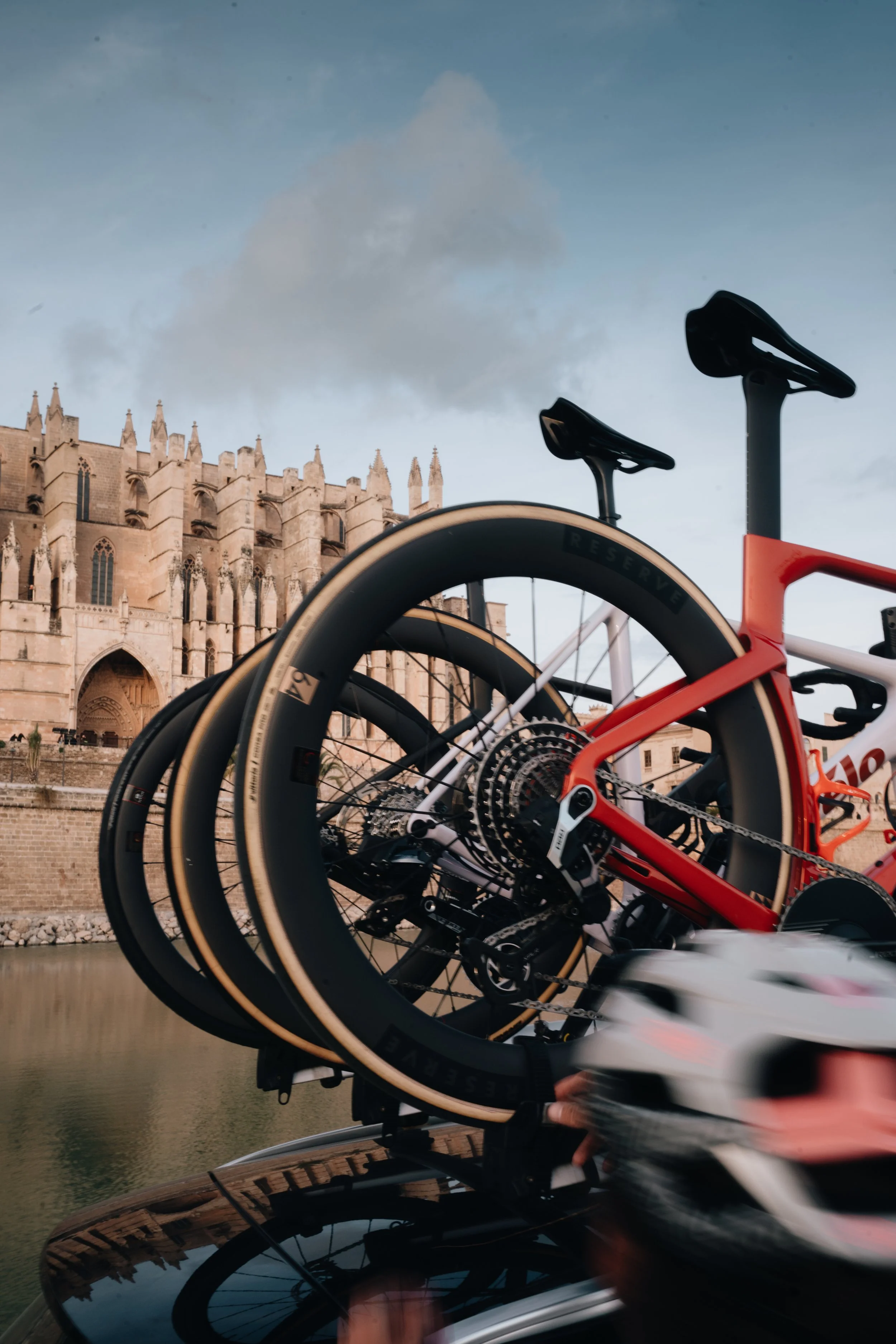 Close-up of a red racing bicycle mounted on a car roof rack, with a white and pink bicycle helmet in motion in the foreground, near a historic stone building, possibly a cathedral, under a cloudy sky.