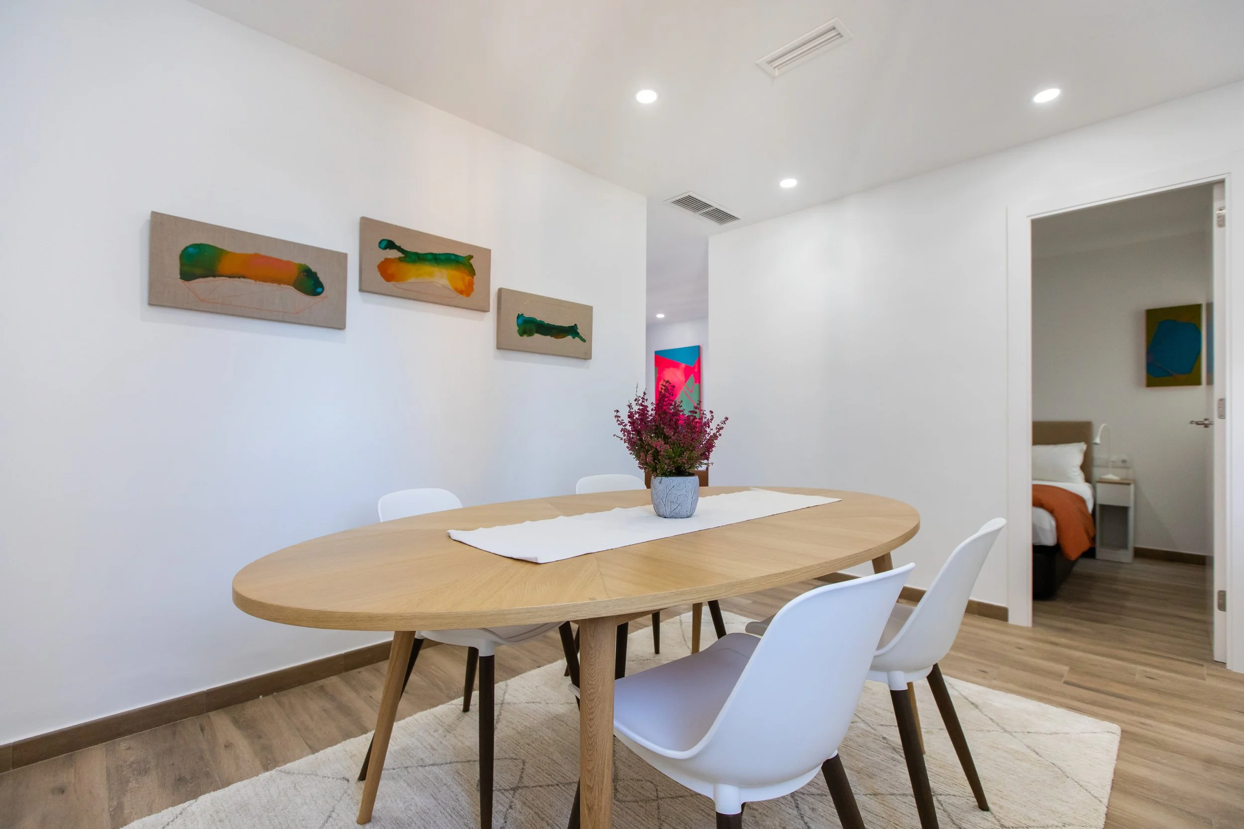 Comedor con mesa de madera ovalada, cuatro sillas blancas y planta de flores moradas en el centro, paredes blancas con arte abstracto, habitación adyacente con cama y cuadros decorativos.