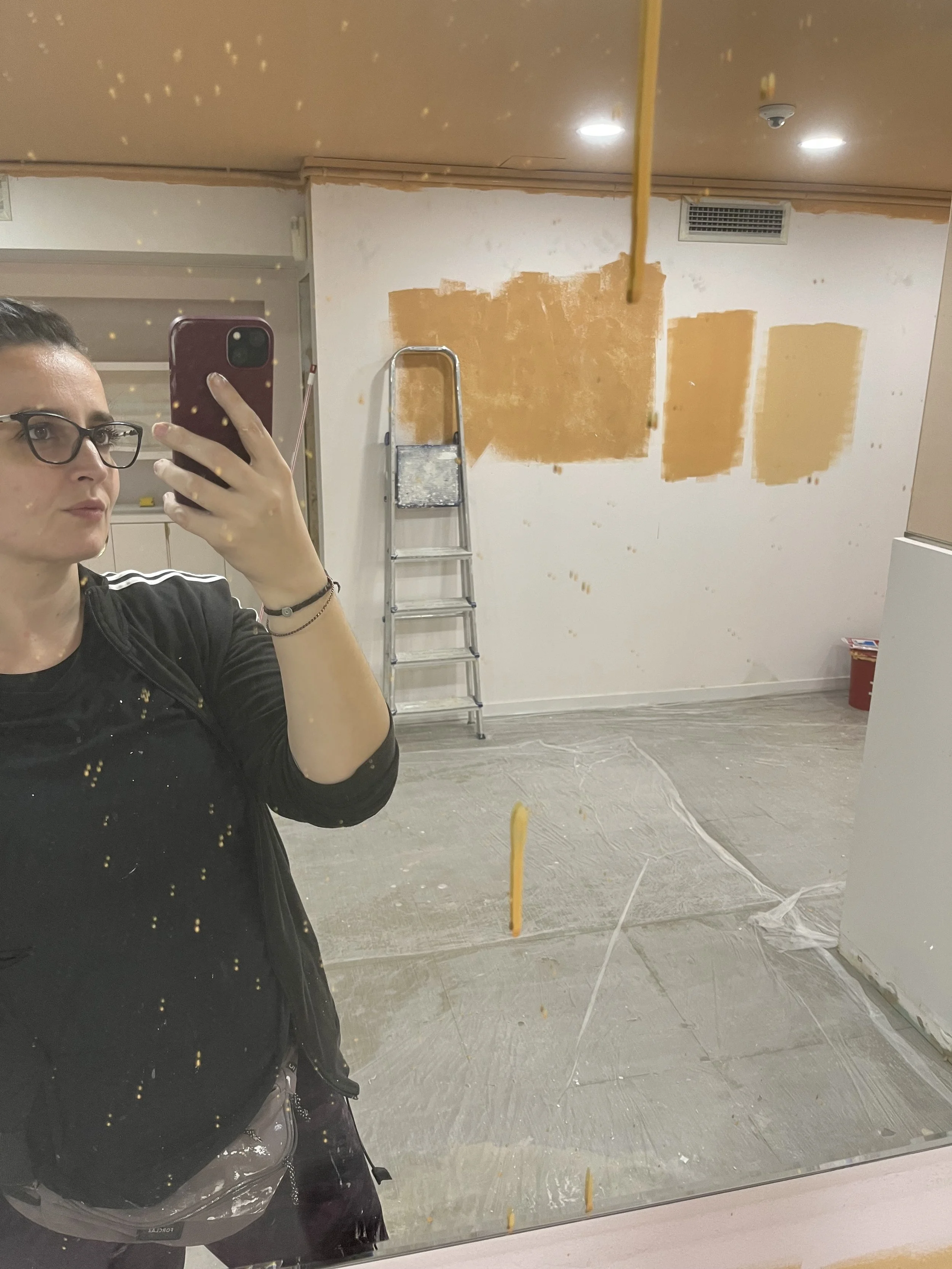 Mujer tomándose una selfie en un espejo en un espacio en construcción o renovación, con paredes parcialmente pintadas en color amarillo y una escalera de aluminio en el fondo.