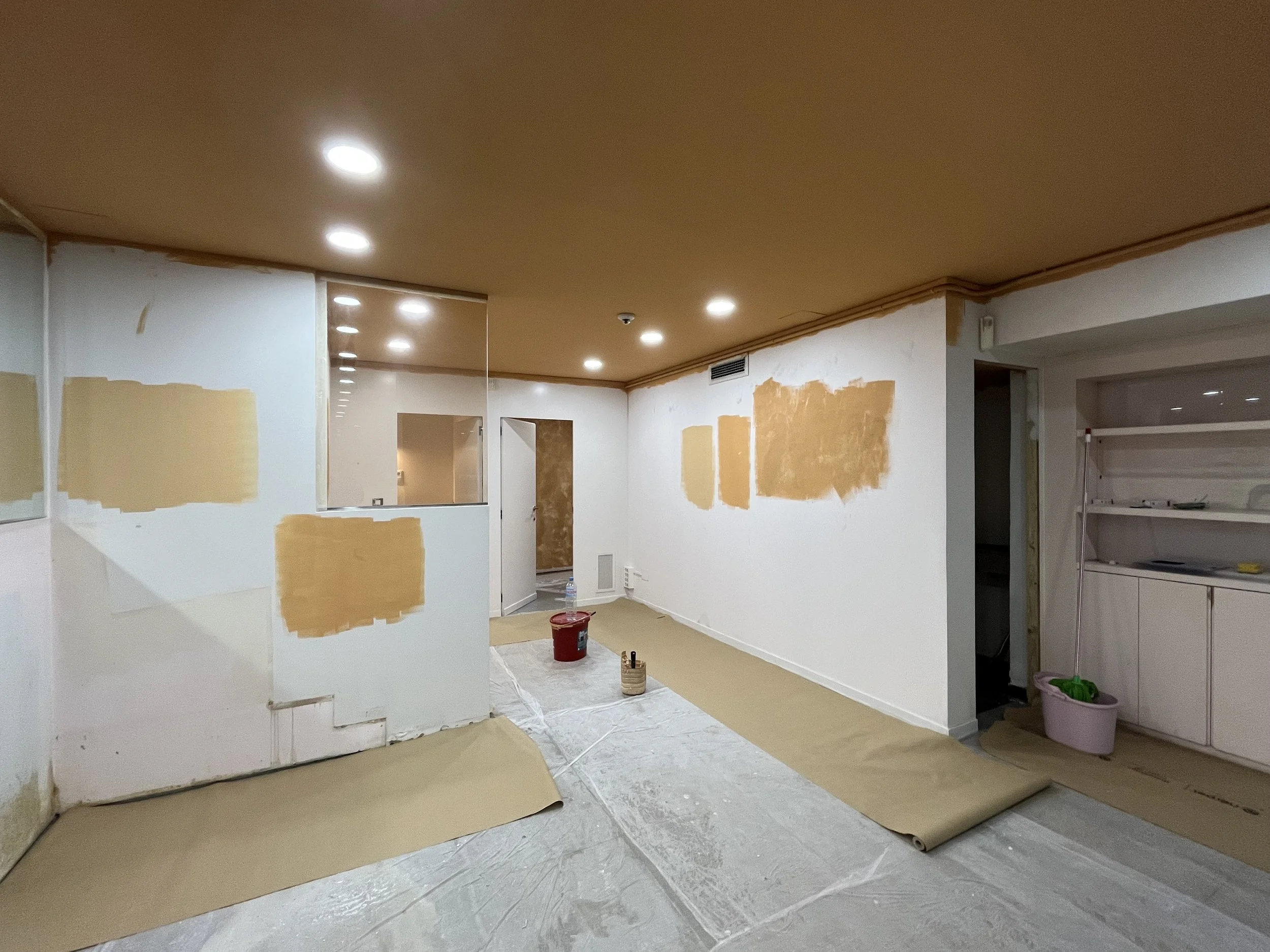 Habitación en proceso de pintura con parches de color beige en las paredes, suelo cubierto con papel protector y varias herramientas de pintura.