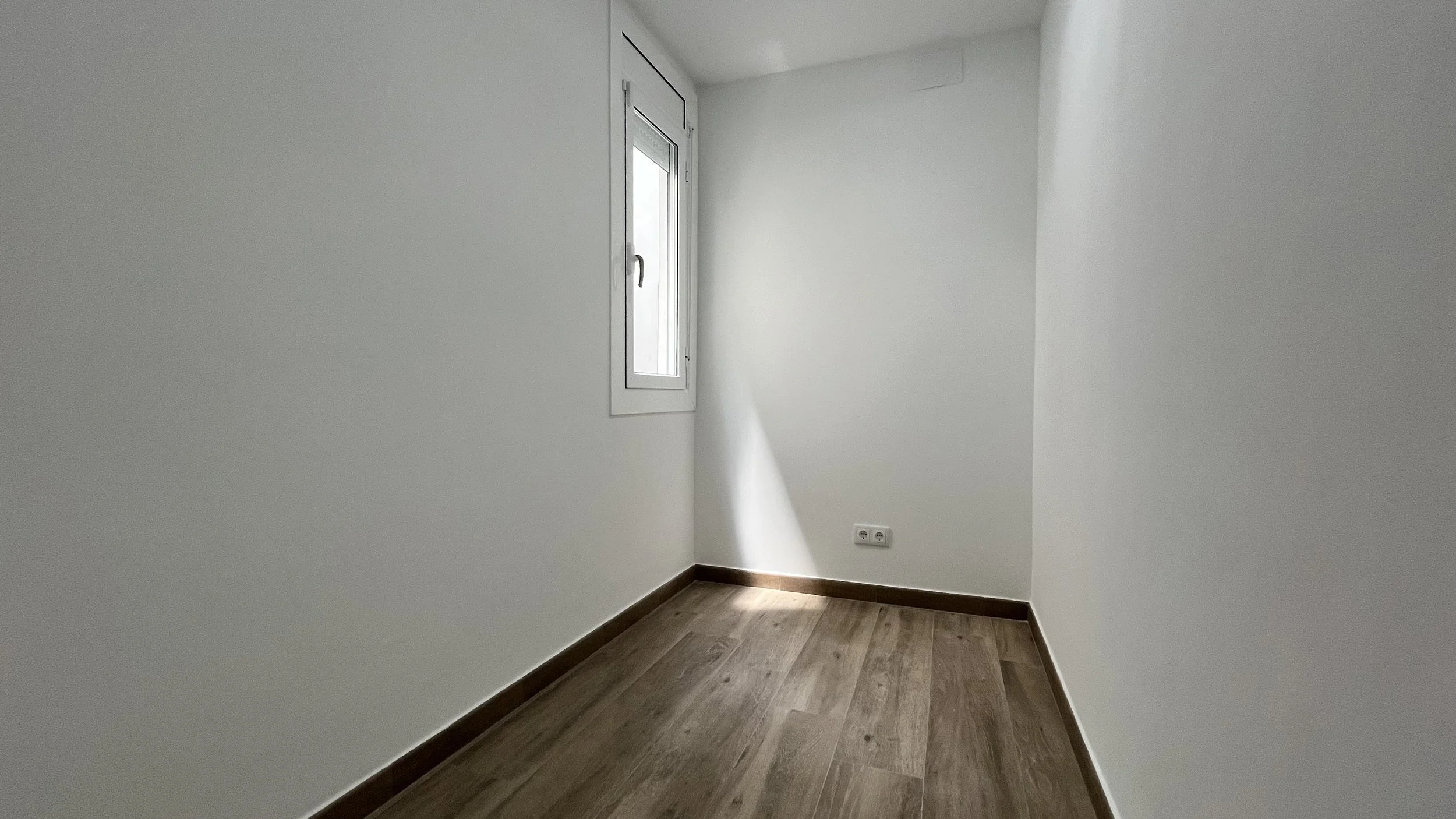 Rincón vacío en una habitación pequeña con paredes blancas, ventana y suelo de madera.