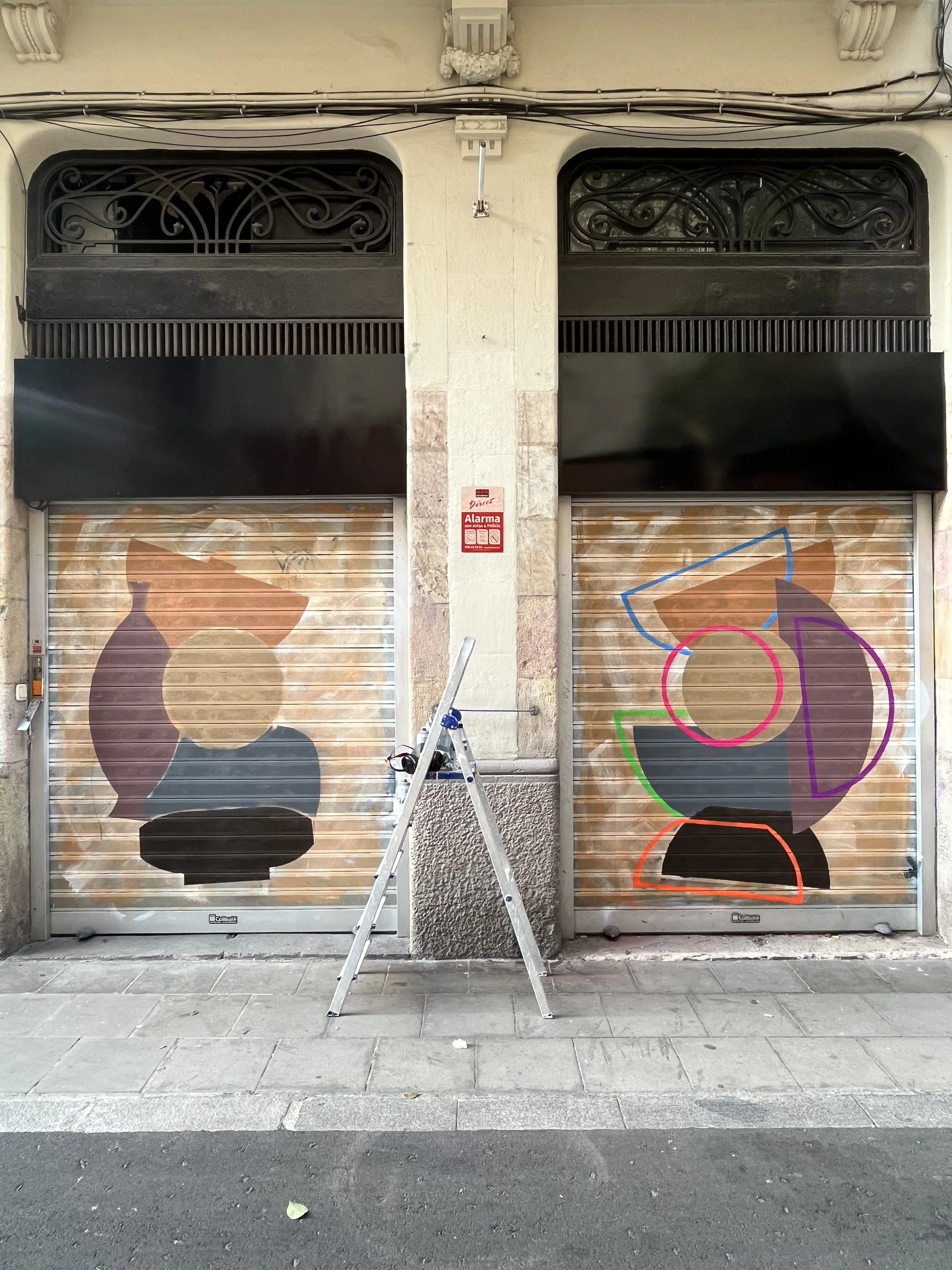 Drehliño de comercio con pintadas coloridas en persianas metálicas cerradas, en un edificio de fachada beige, con escalera y señal de alarma en el centro.
