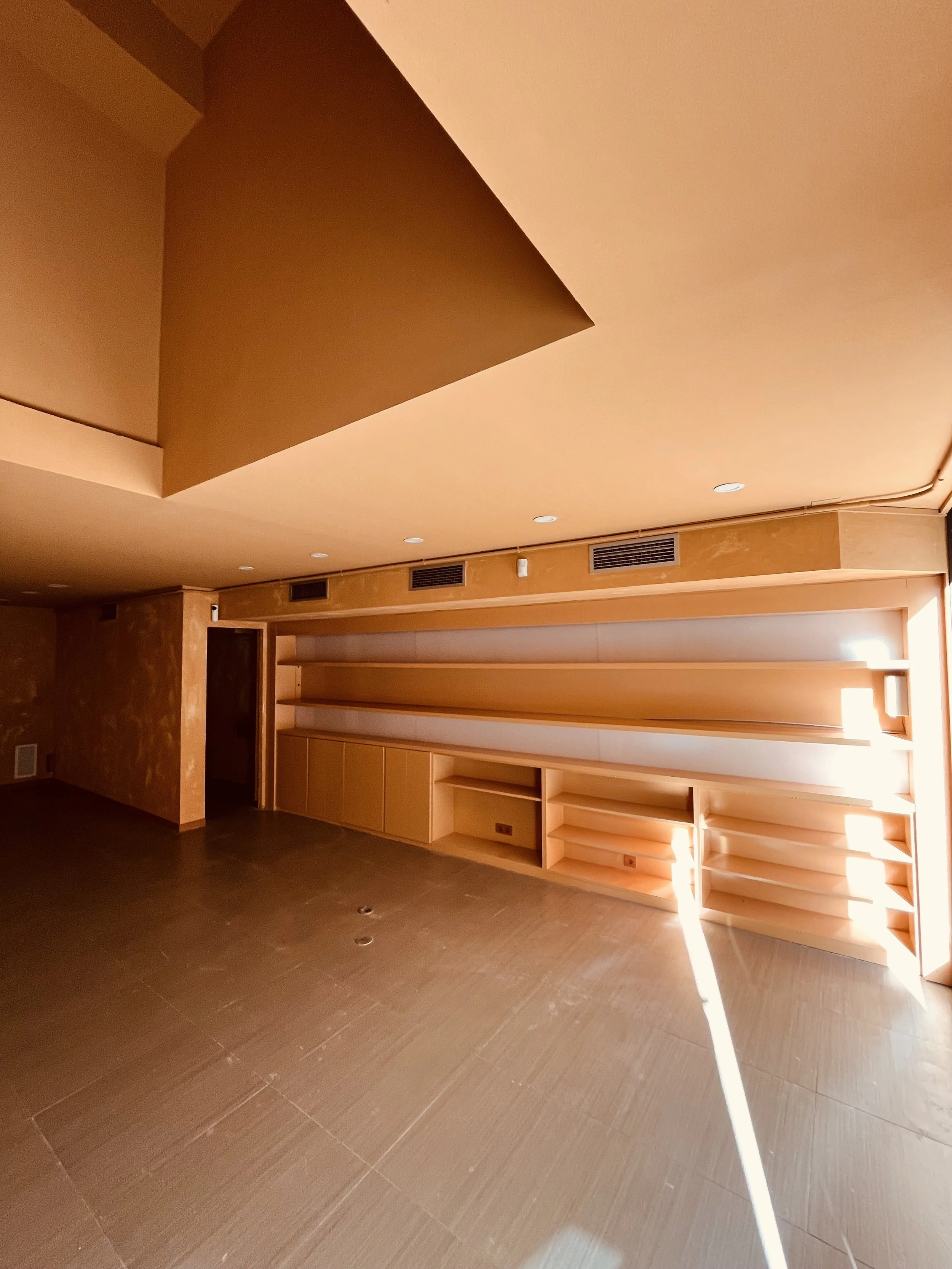 Sala vacía con estanterías de madera a la derecha y paredes en tonos anaranjados, con ventanas por donde entra luz solar.
