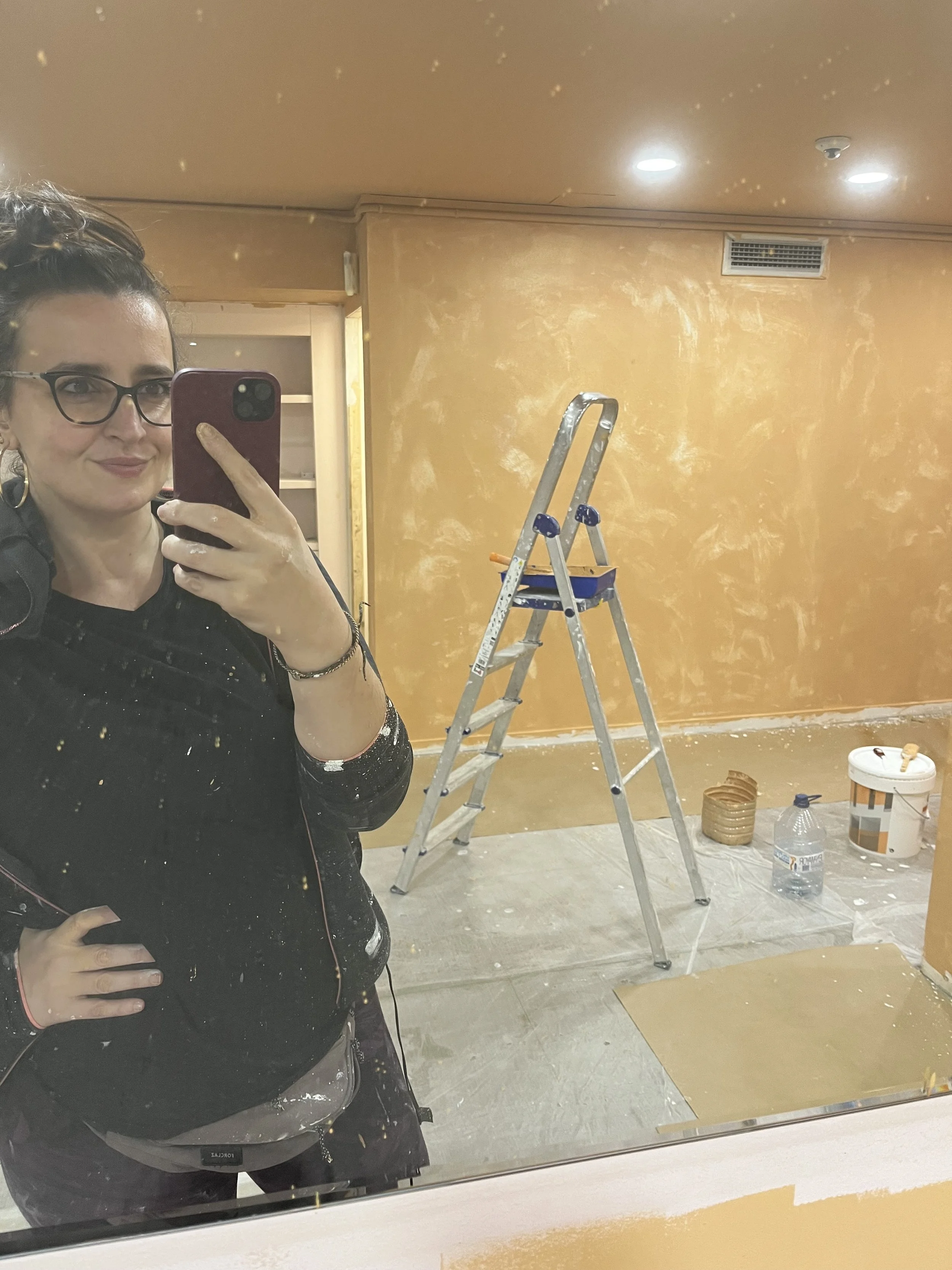 Una mujer tomándose una selfie en un espejo en una habitación en proceso de pintado, con una escalera de aluminio, baldes de pintura y una botella de agua en el suelo.