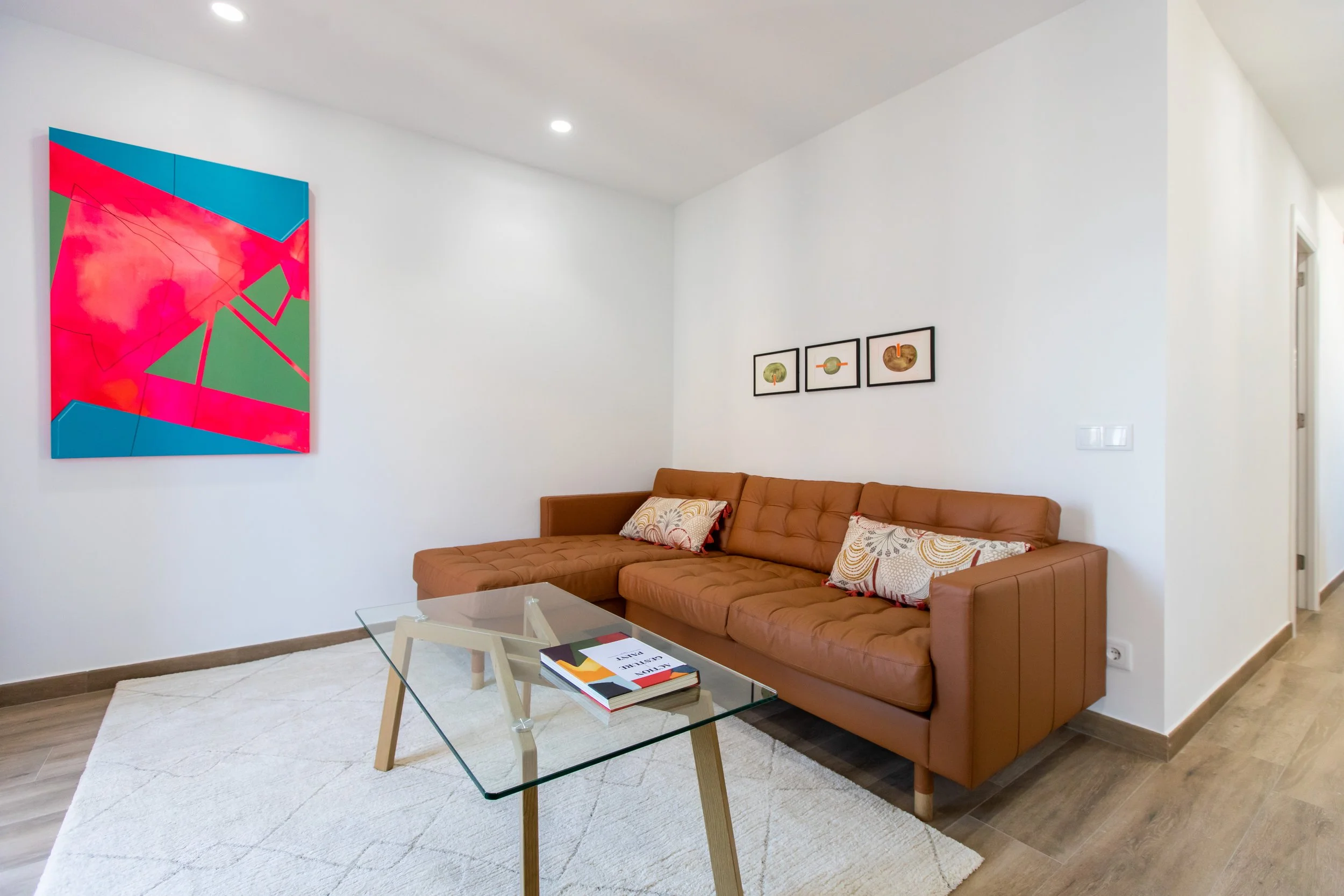 Sala de estar con sofá de cuero marrón, mesa de vidrio, cuadros abstractos en la pared y pintura colorida en la pared opuesta.