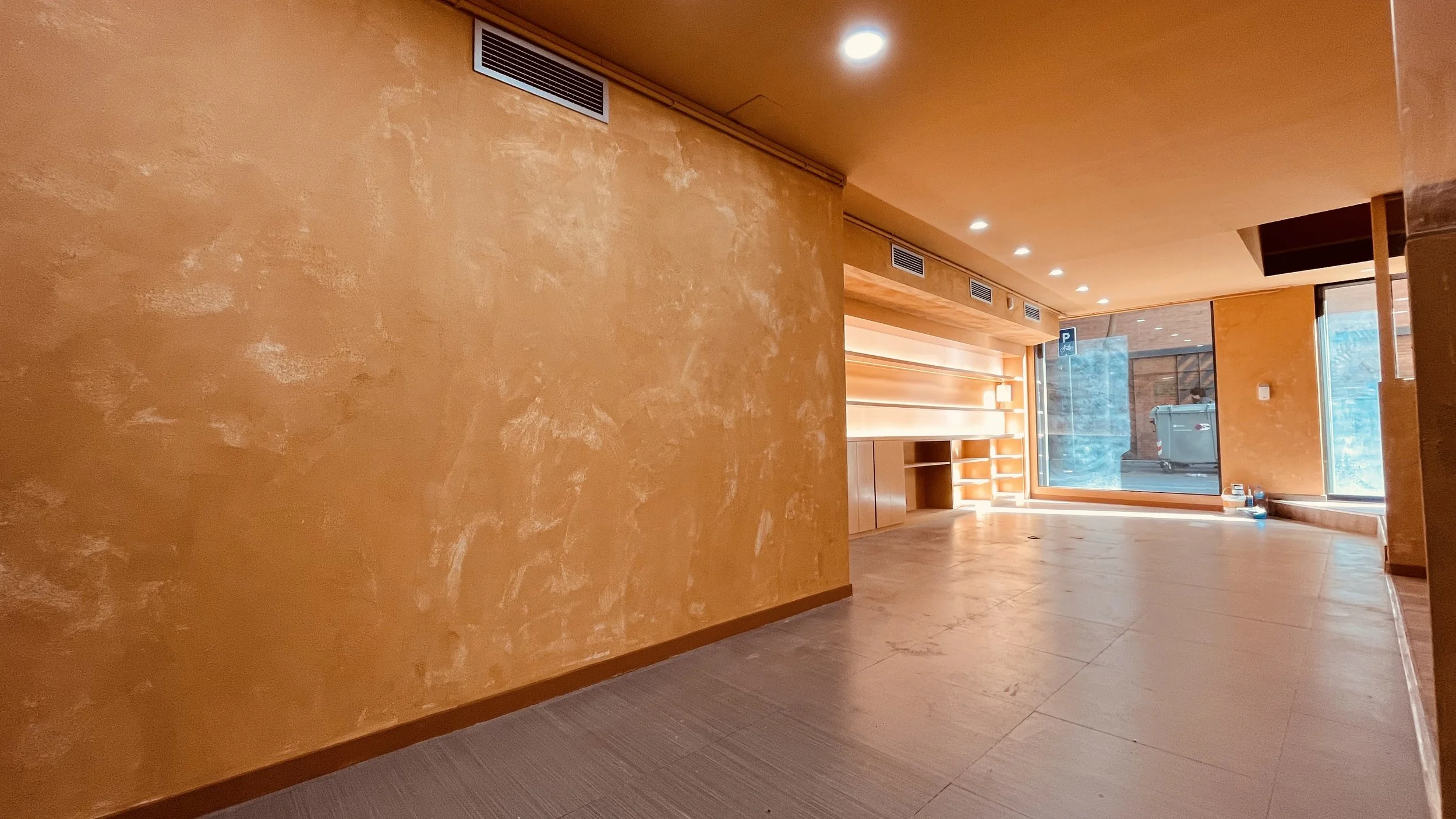 Espacio interior con paredes naranjas y suelos de color marrón, con ventanas grandes que permiten la entrada de luz natural, y una pequeña área de almacenamiento o exhibición en la pared