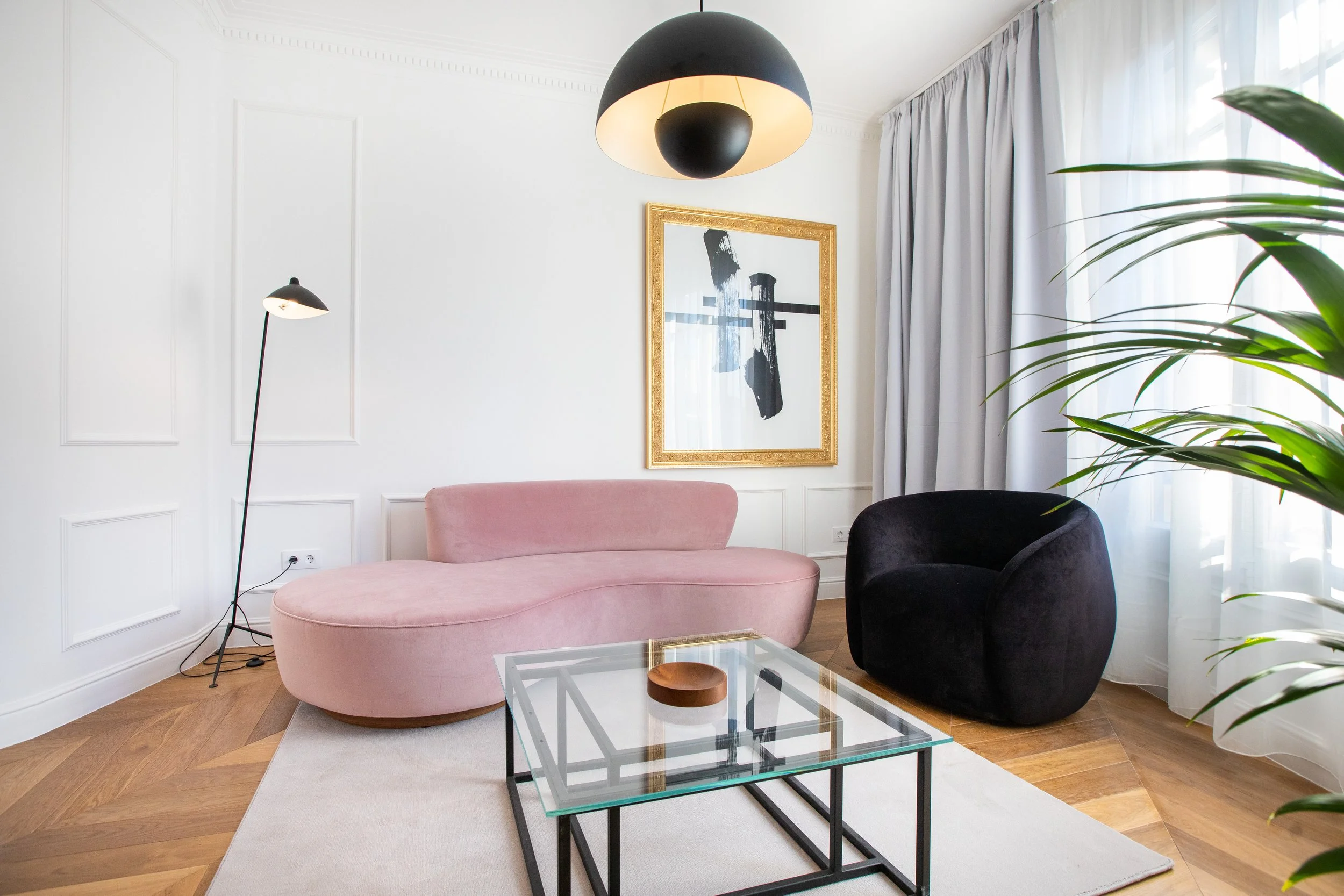 Sala de estar moderna con sillón rosa, sillón negro, mesa de cristal y cuadro abstracto con marco dorado, iluminada por lámpara de techo negra y lámpara de pie, con cortinas blancas y planta verde.