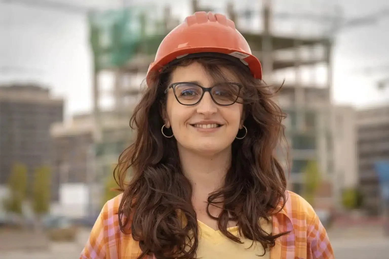 Mujer con gafas y cabello rizado, usando casco de construcción naranja, sonriente, en un entorno de obra o construcción.