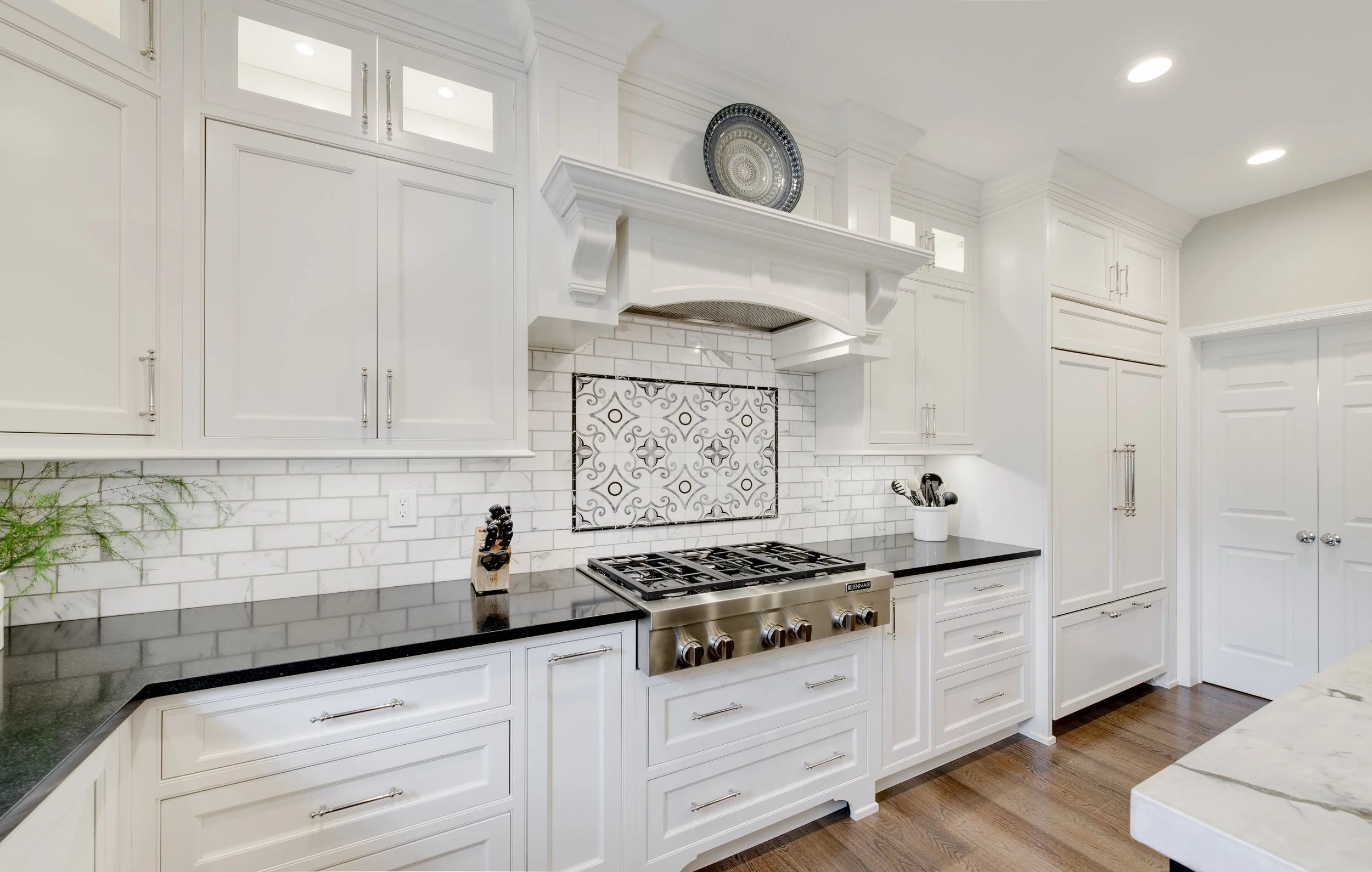 Modern white kitchen with black countertops, stainless steel stove, decorative tile backsplash, white cabinetry, and hardwood floor.