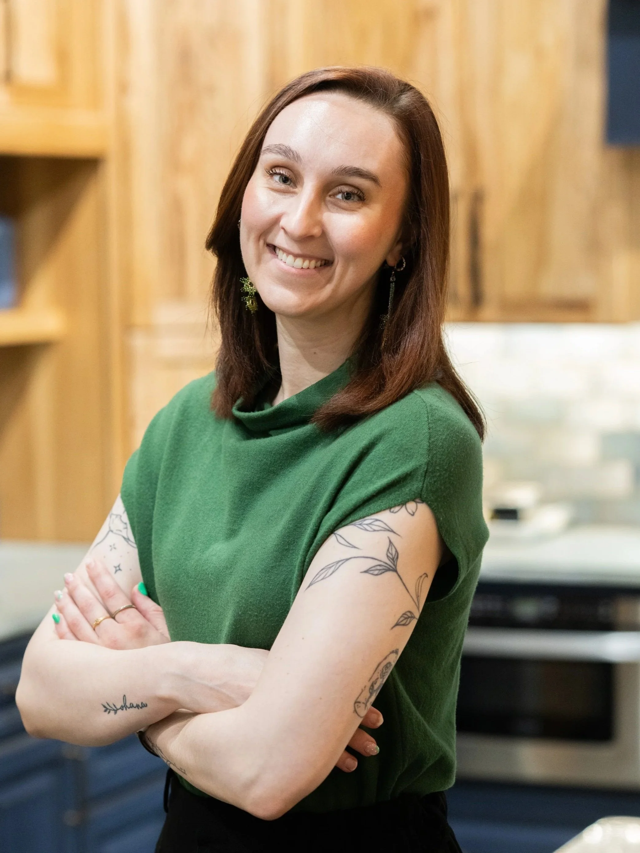 A woman with dark brown hair, wearing a green top, smiling with arms crossed, standing in a room with wooden and dark-colored furniture.