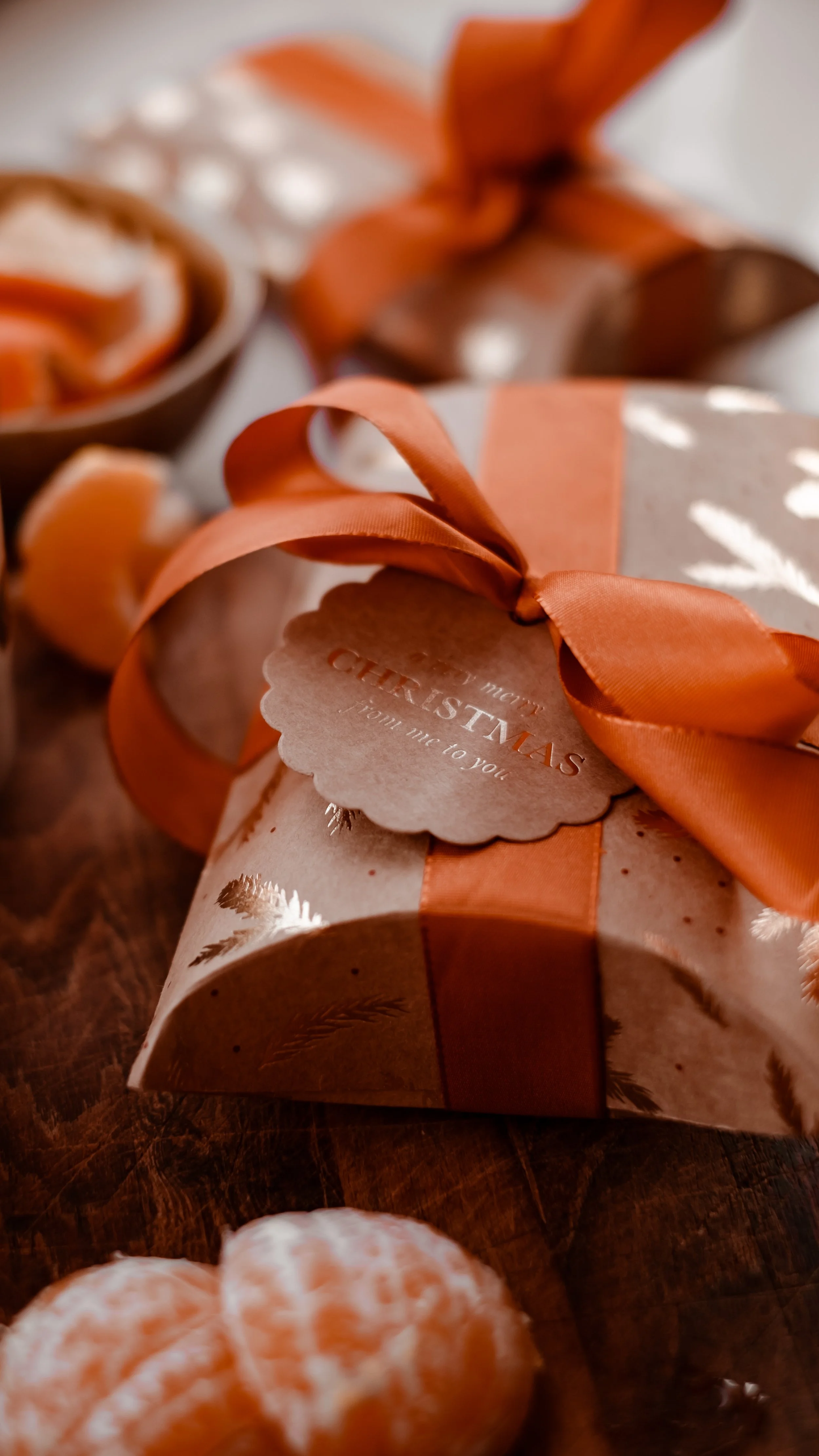 Julegave med brun indpakning, orange bånd og julefremme, ligger på træbord.