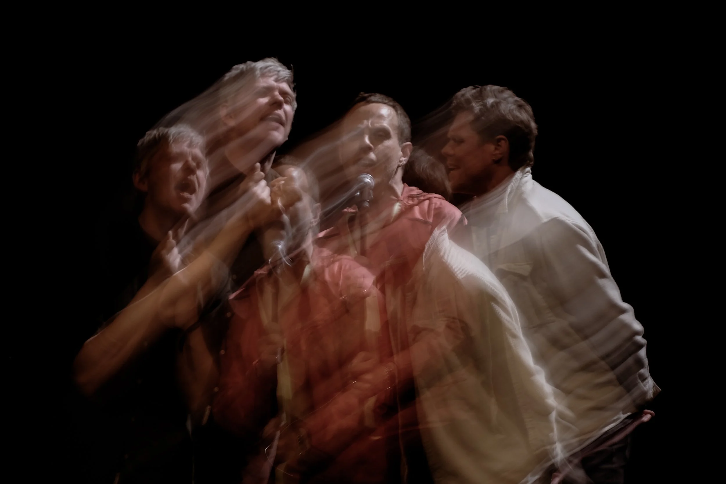 Billed af fire personer, der synger og optræder på en scene med lys, og der er en bevægelseffekt, der skaber en sløredeudseende.