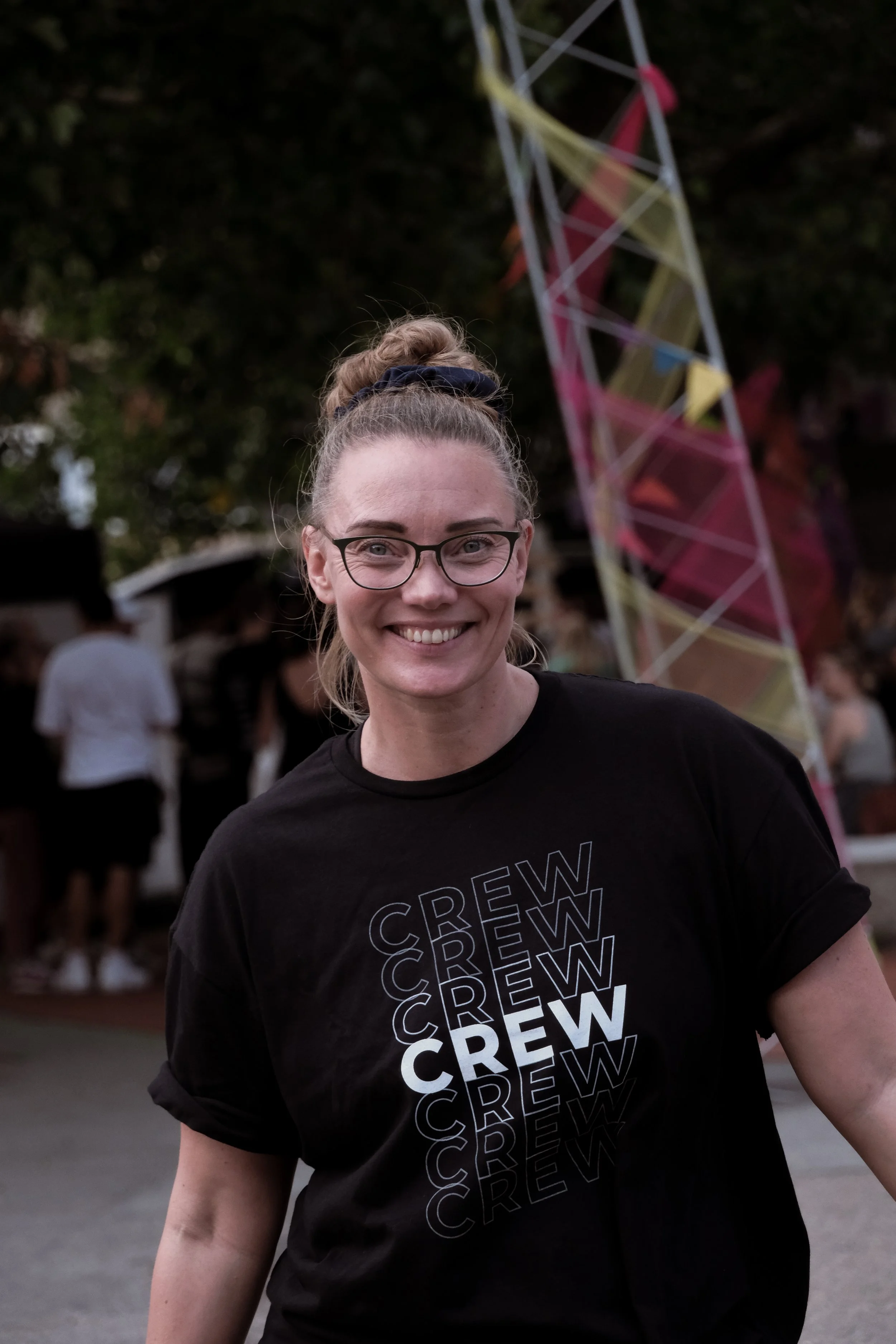 Smilende kvinde med briller, trøje med teksten 'CREW' i flere lag, udendørs arrangement med mennesker og farverige dekorationer.