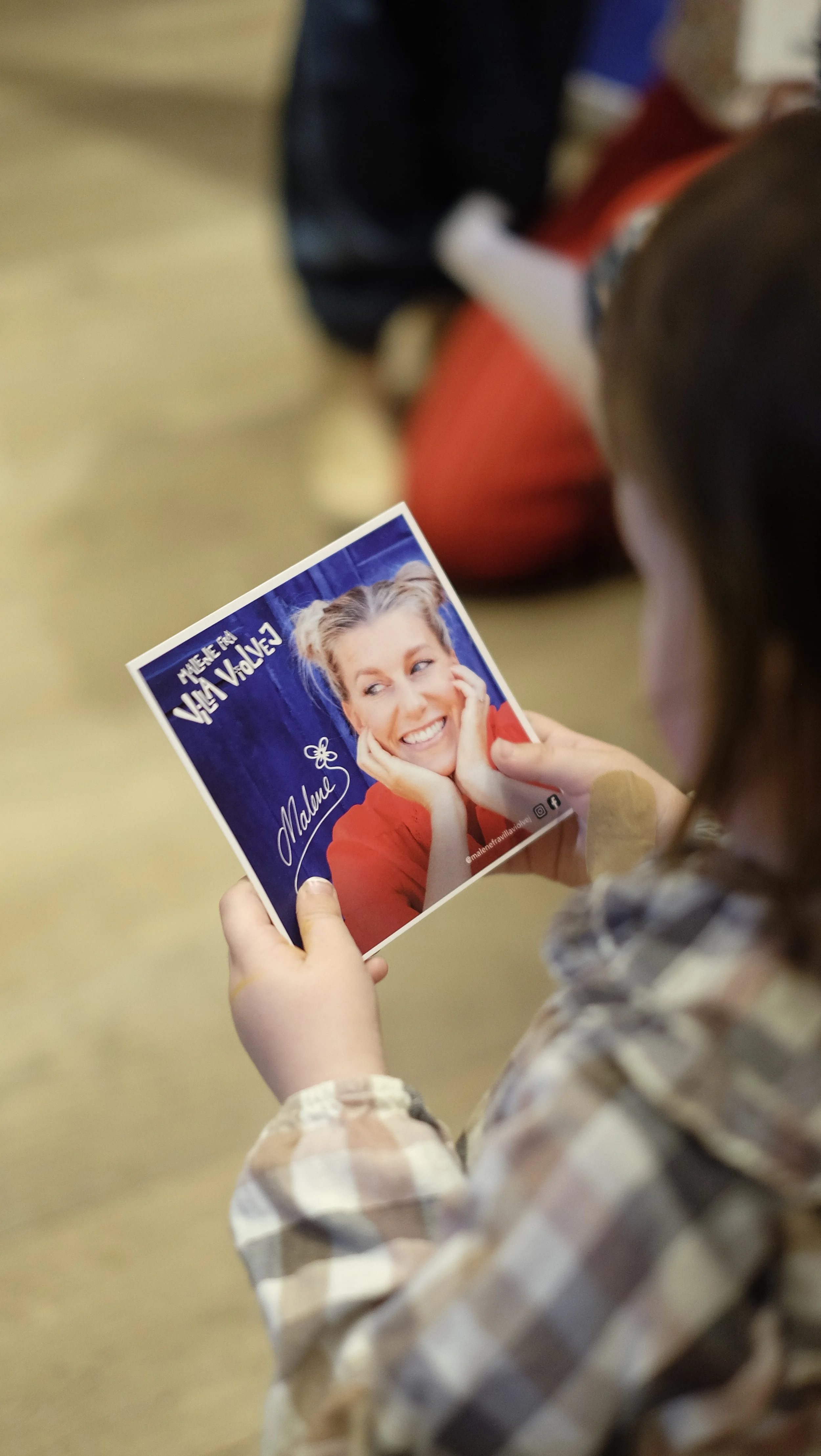 En person holder et foto med en smilende kvinde i en rød top, der er signerede. Fotoet har tekst på dansk og sociale medie ikoner.