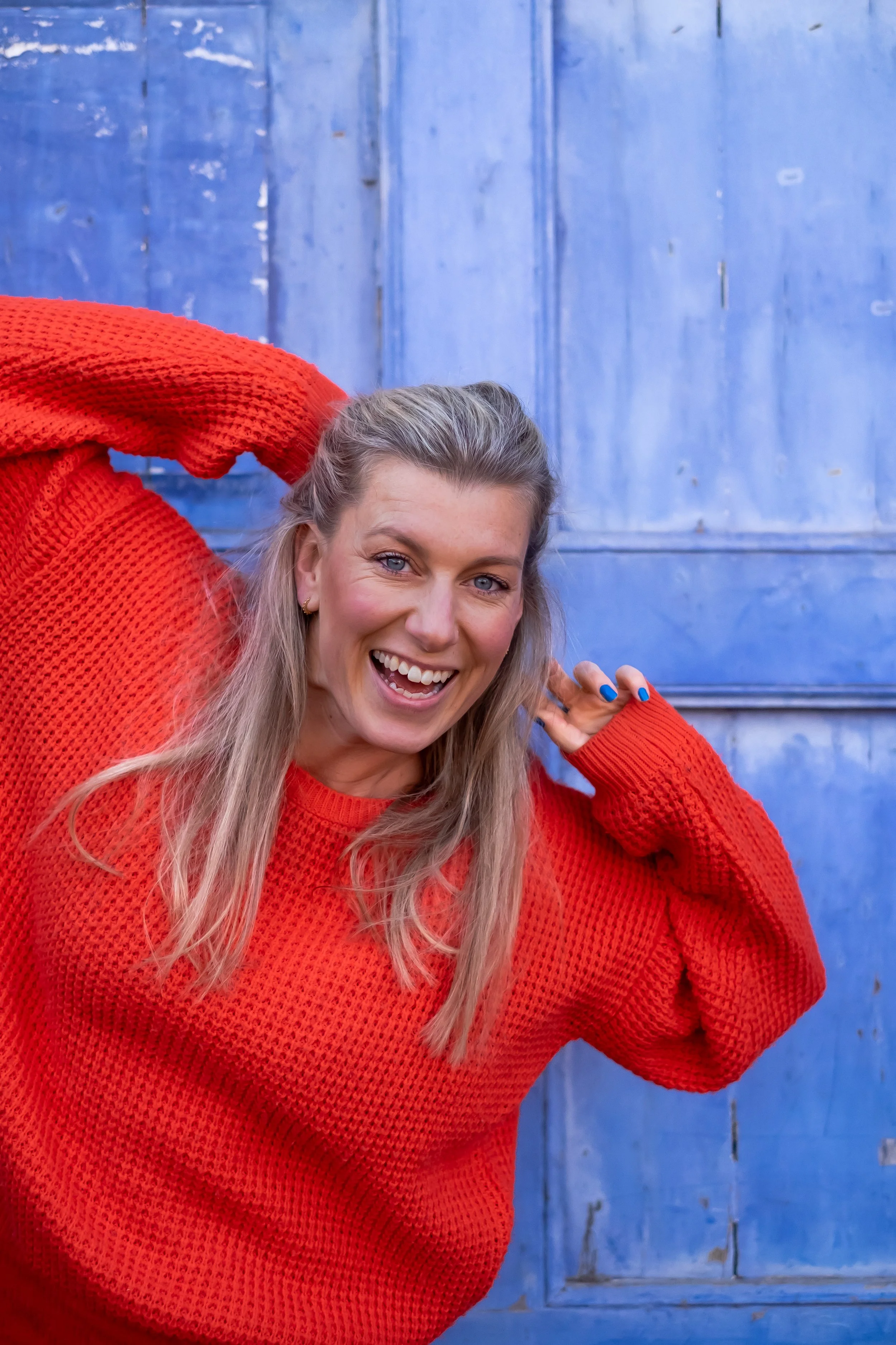 En glad kvinde med blondt hår iført en rød strikket sweater, der står foran en blå trævæg og smiler til kameraet.