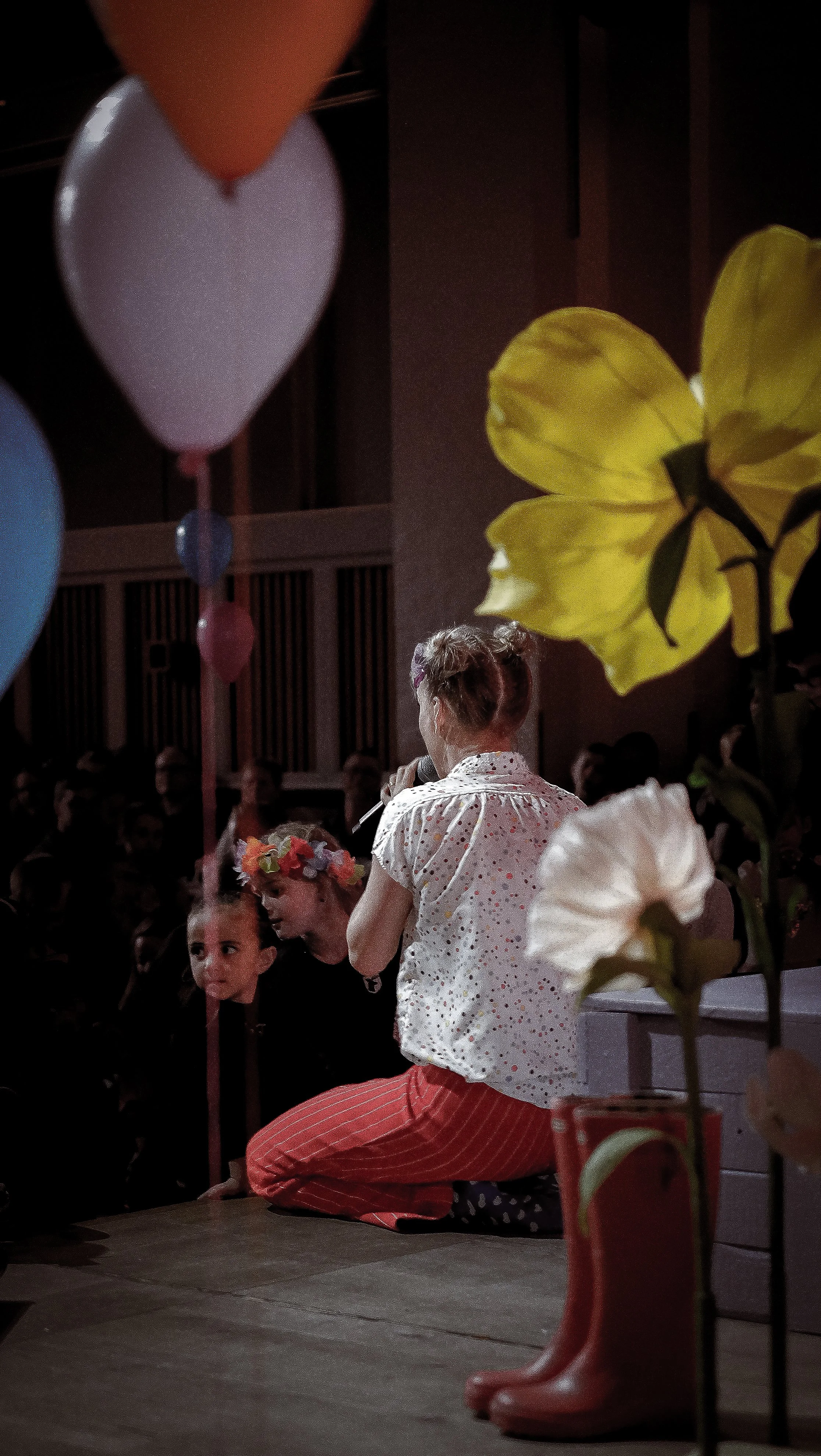 En kvinde taler til en gruppe børn, med balloner og blomster i forgrunden, i et mørkt lokale.