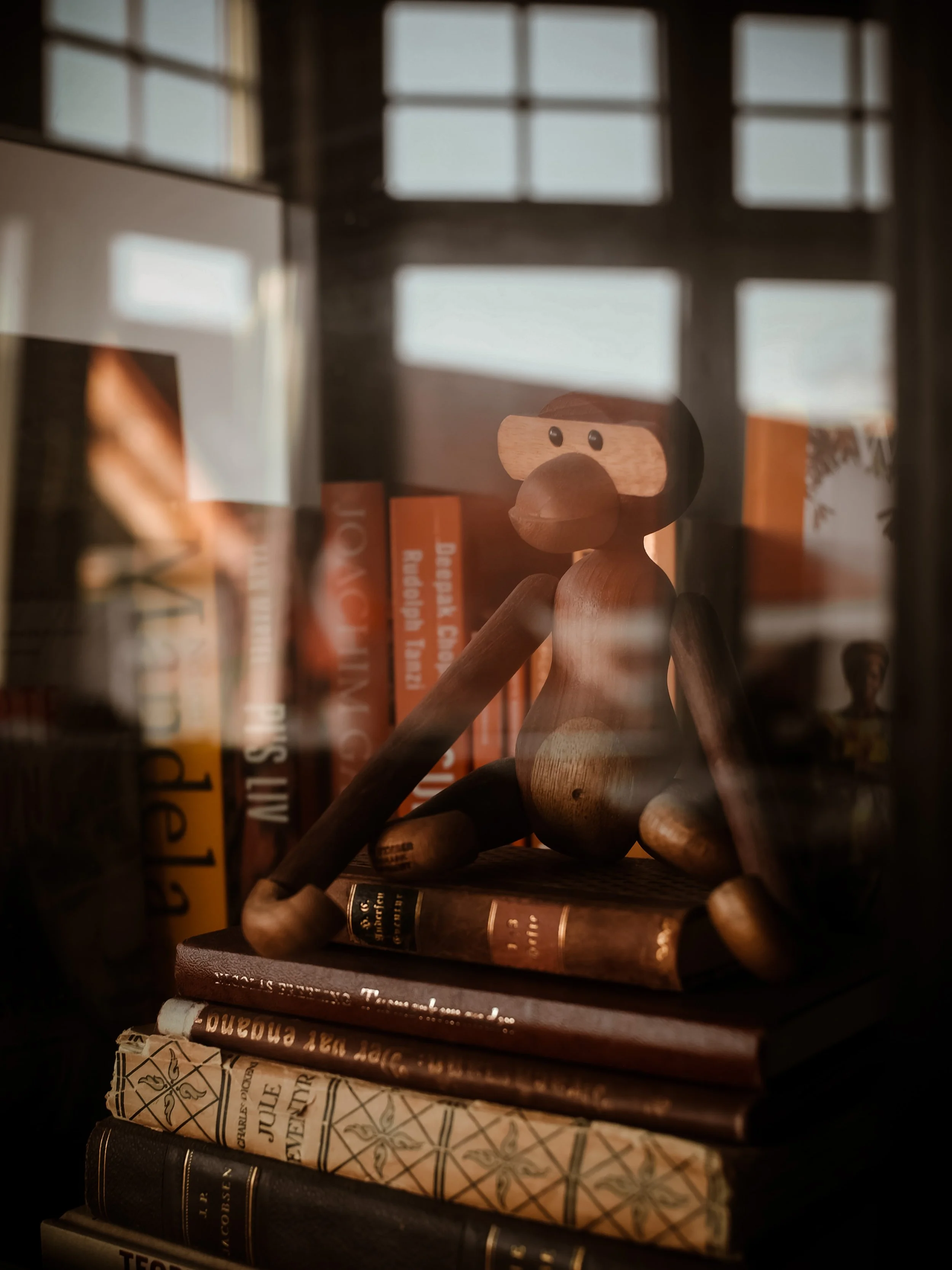 En træ dukke med store øjne sidder på en stak af bøger bag et vindue.