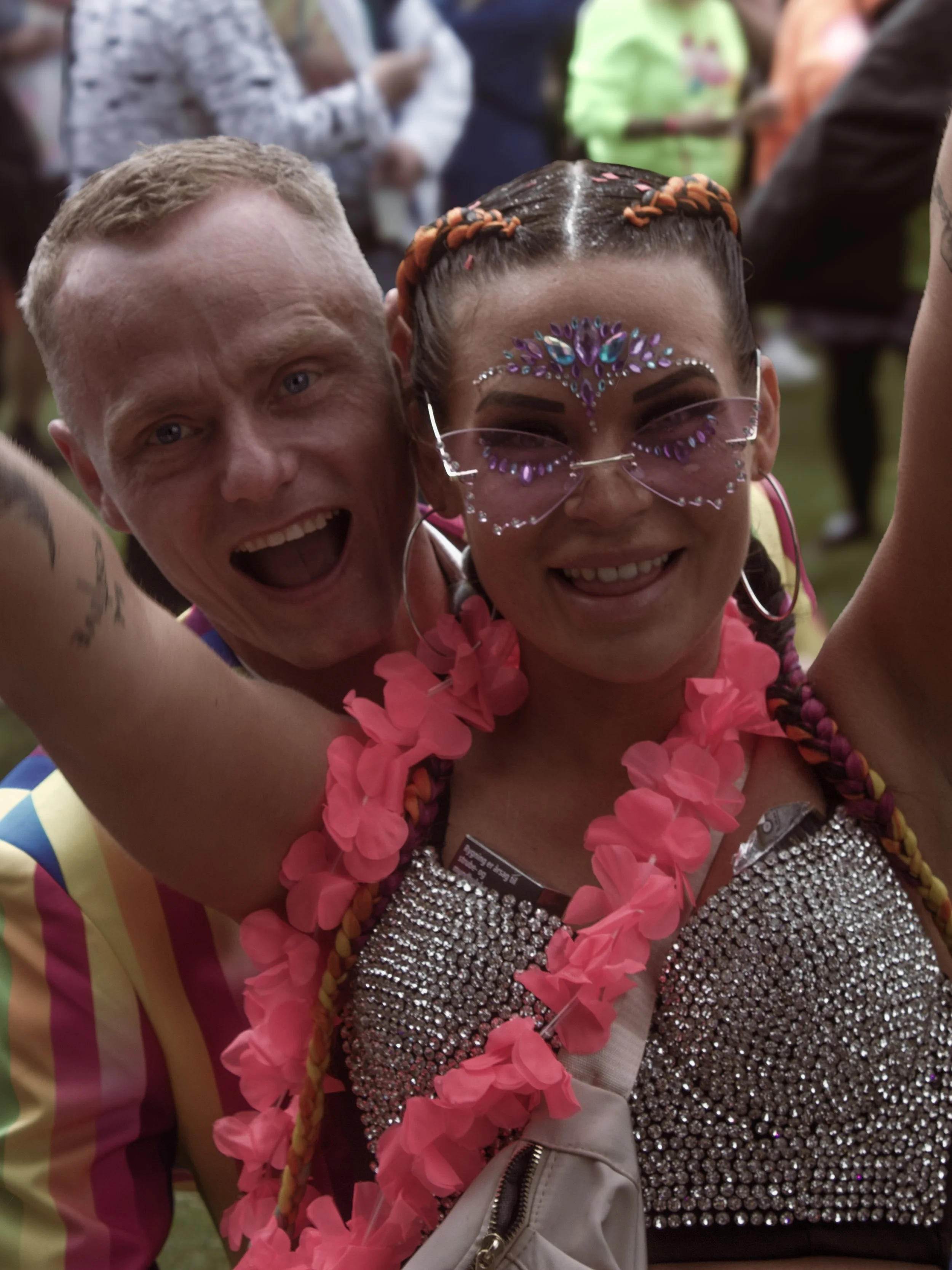 To mennesker deler et sjovt øjeblik, den ene med blondt hår og den anden med mørkt hår, iført farverigt og dekorativt tøj, omgivet af en gruppe mennesker udendørs.