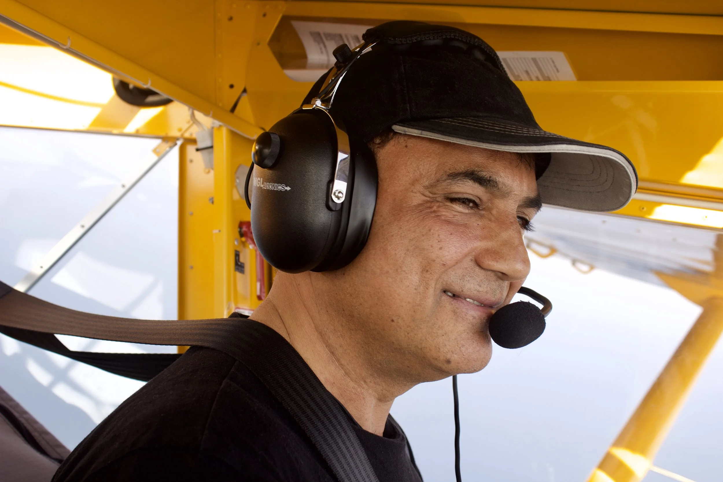 Mand i flyvemaskine med høreværn og mikrofon, flyver i luftet.