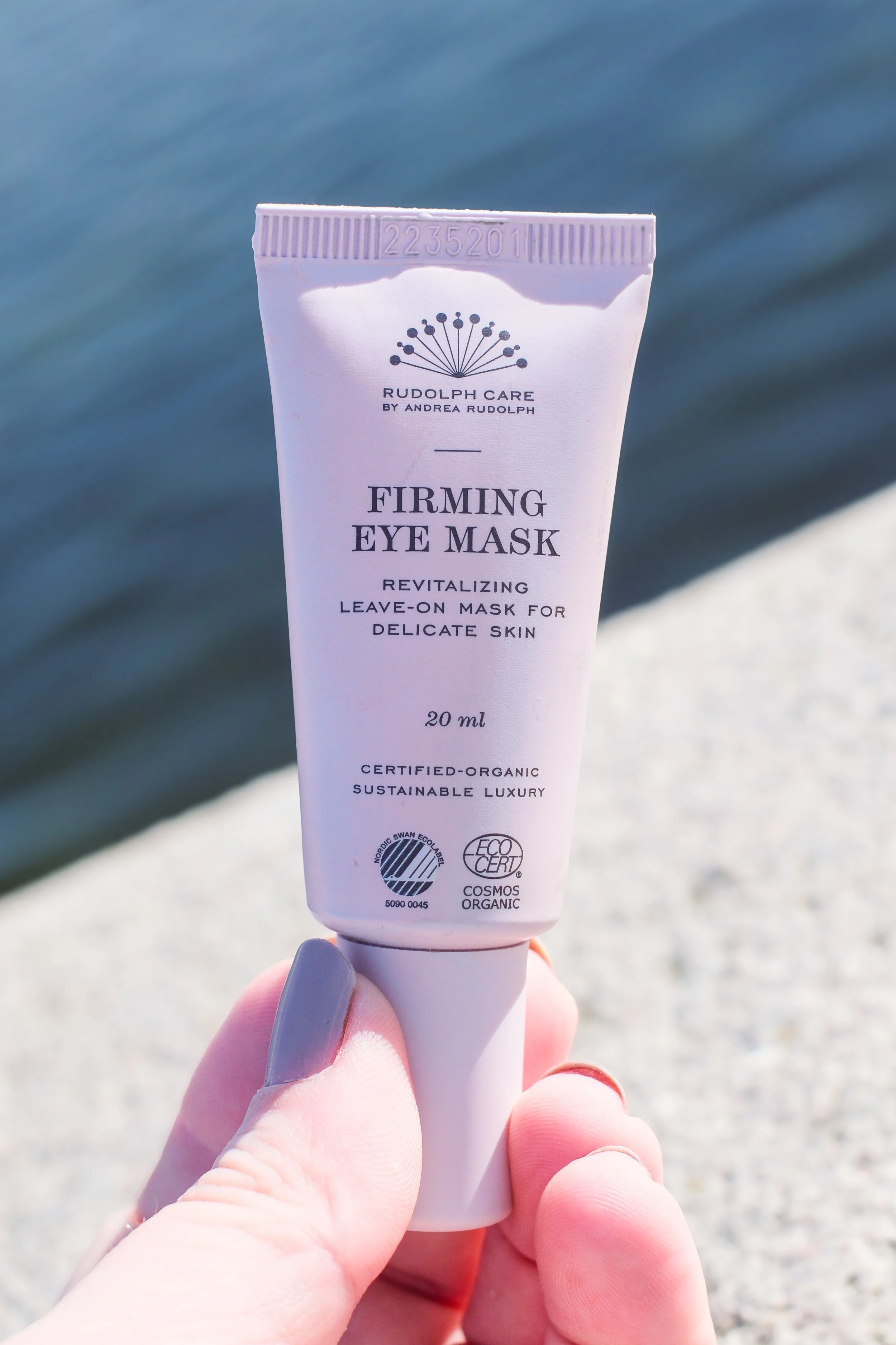 Hånd holder en hvid tubetag med ansigtsskygge bag vand, der indeholder Rudolf Care firming eye mask.