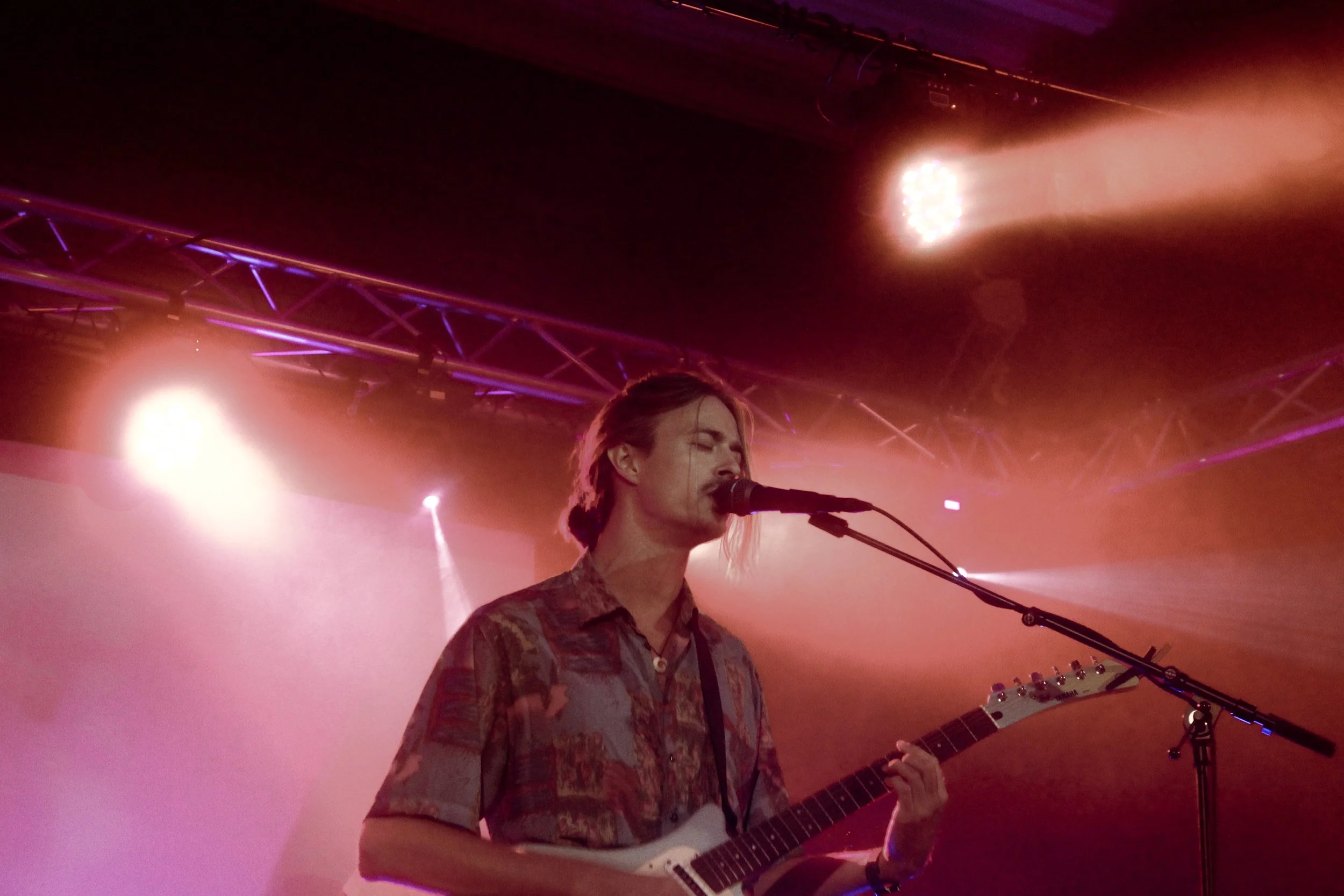 Musiker spiller på scenen med guitar og mikrofon under koncert, med lyssætning i baggrunden.