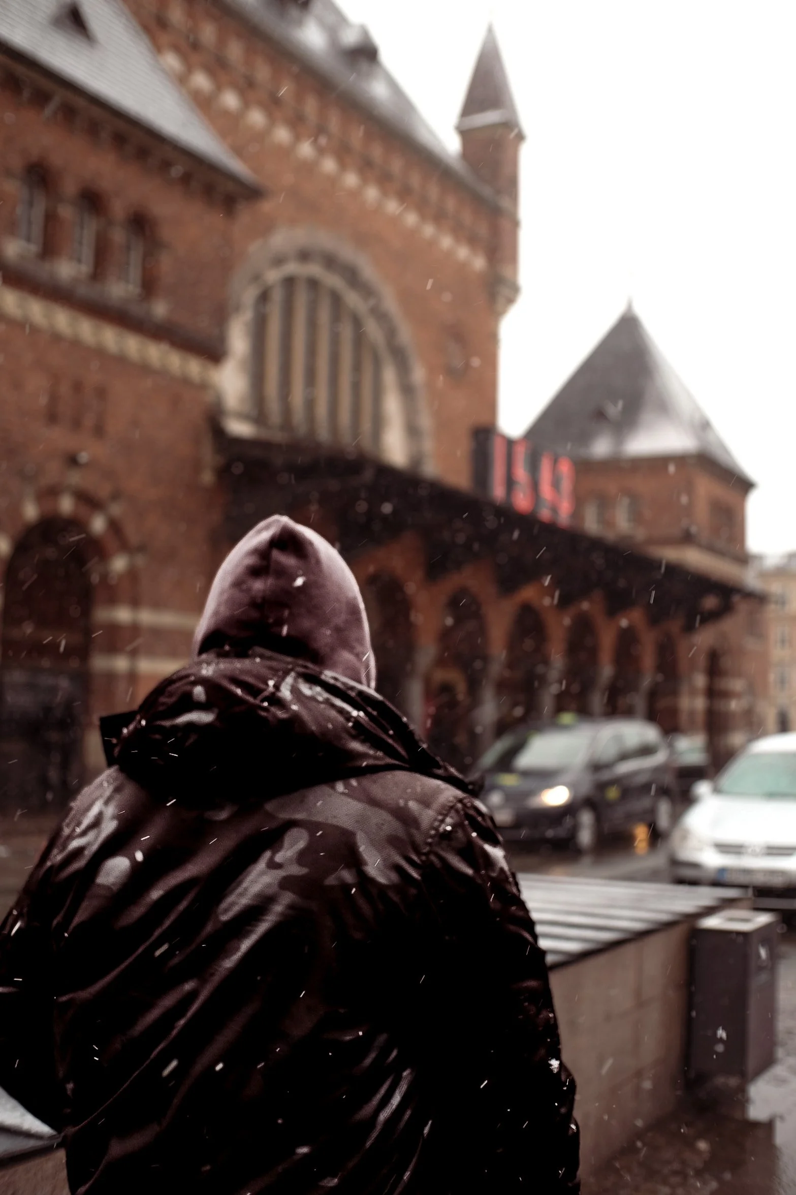 Person med mørk jakke og hættetrøje, set fra bag, står foran en rød murstensbygning i regnvejr.