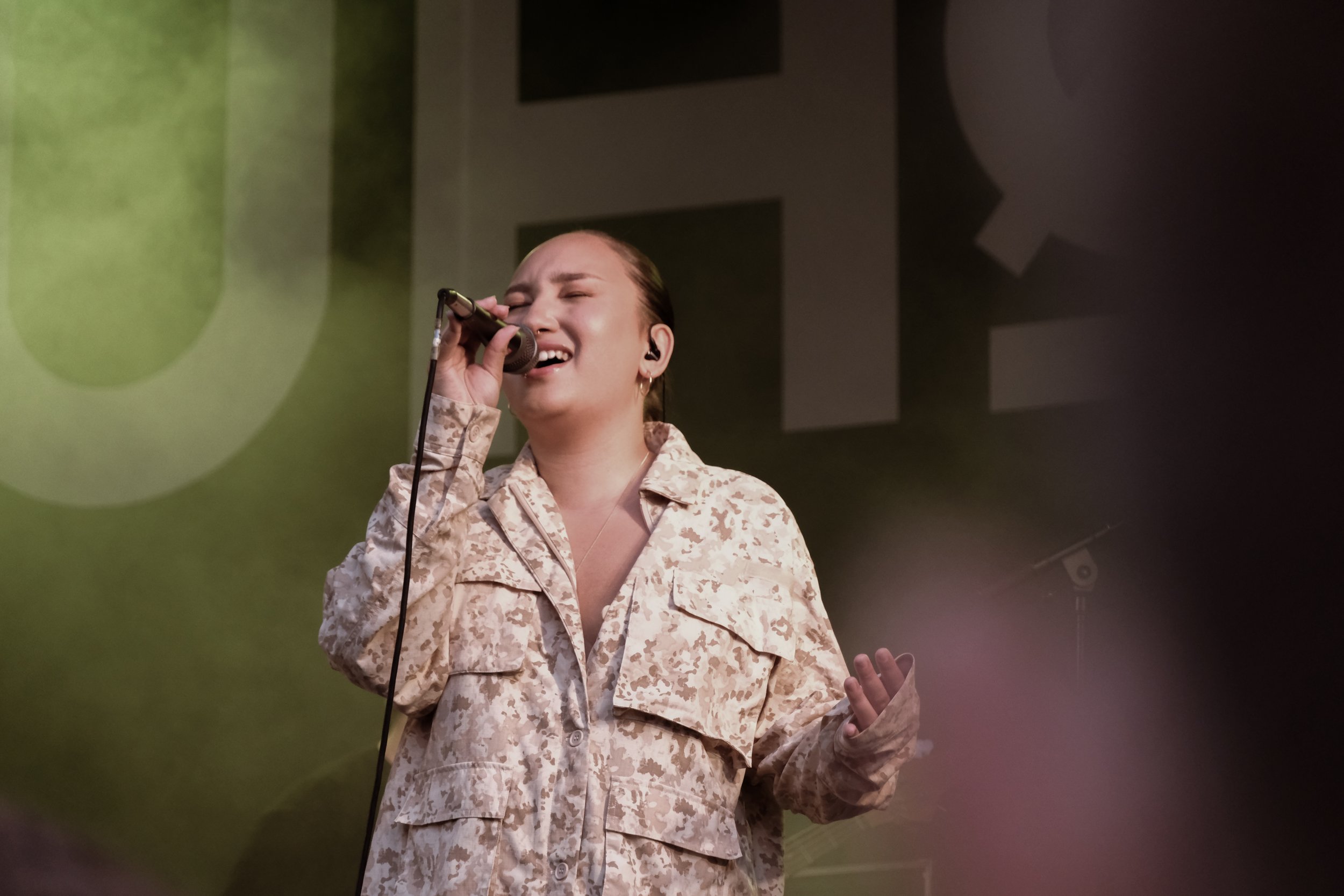 En kvinde synger med mikrofon på en scene, iført militærinspireret tøj i beige camouflagemønster.